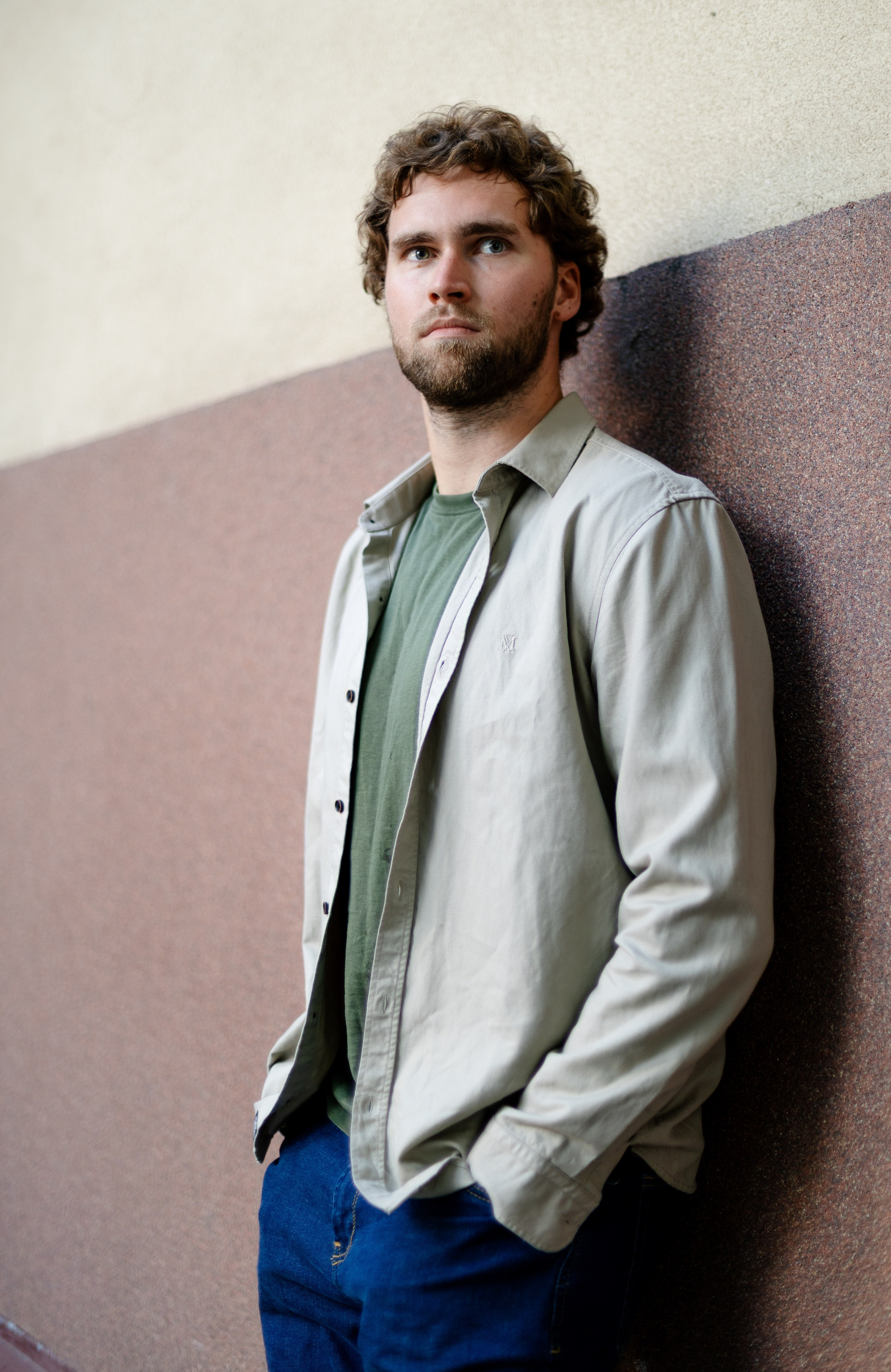 A relaxed full-length street portrait of a curly-haired man leaning against an urban wall in soft natural light