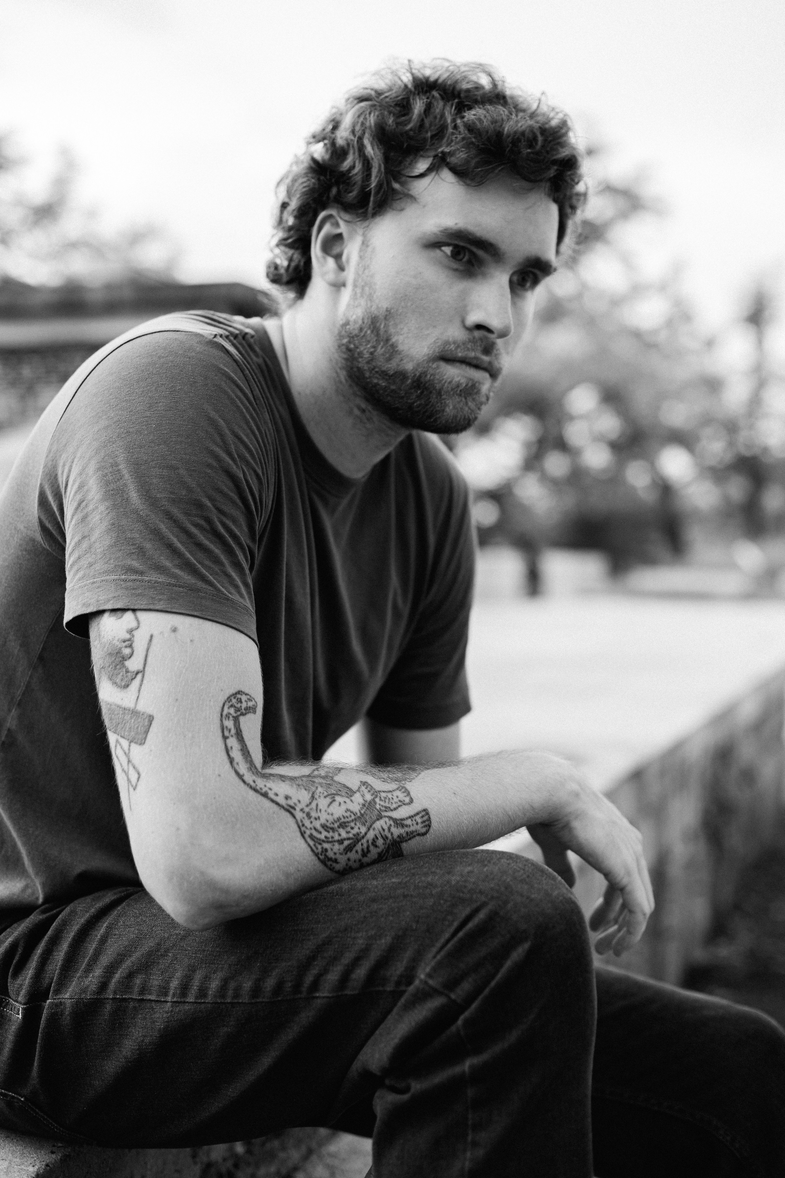 A black and white street portrait of a man with tattoos sitting outdoors looking away from the camera