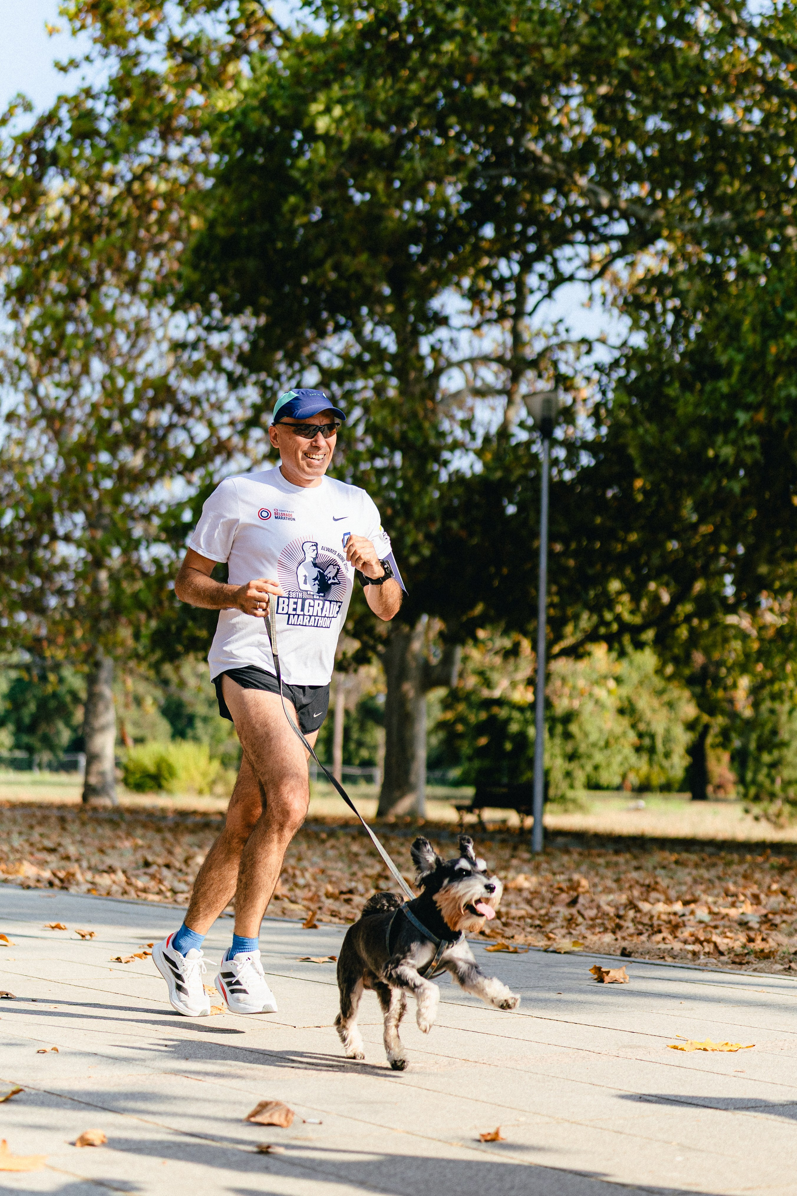 A smiling man running in Belgrade park wearing a "Belgrade Marathon" t-shirt, leading a small schnauzer dog on a leash during the S95 community run.