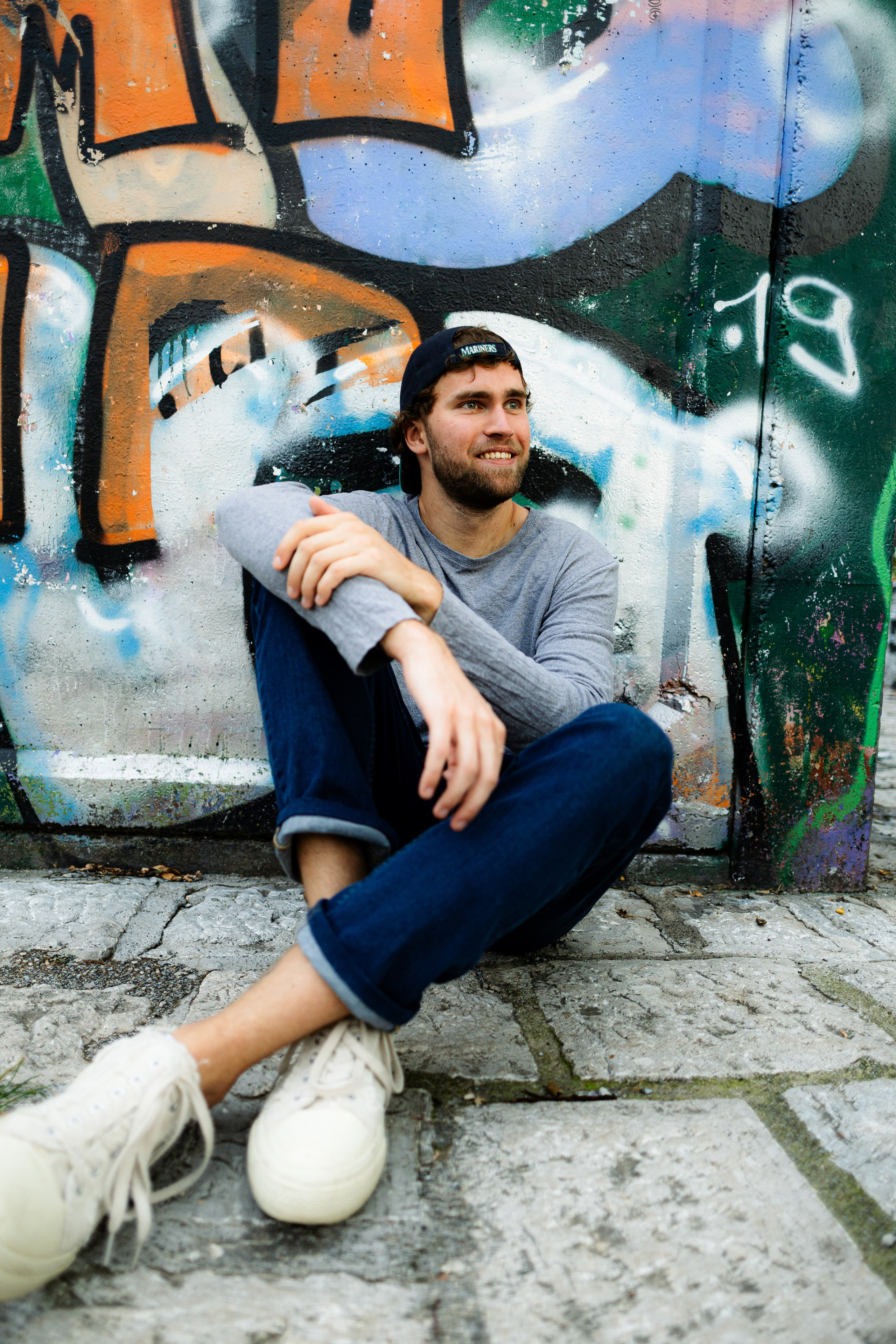 A casual young man with curly hair and a cap sitting relaxed on a sidewalk near a graffiti wall