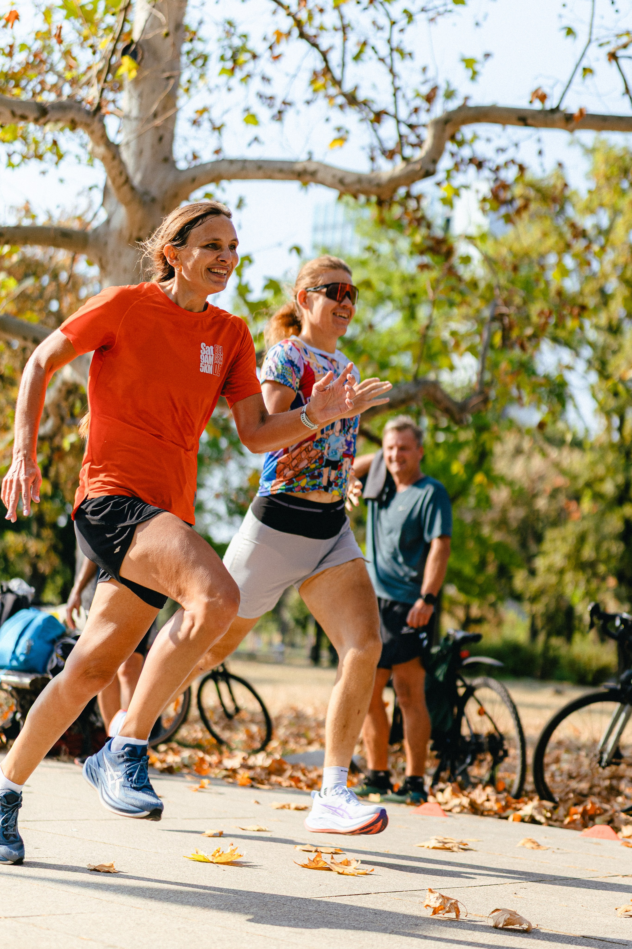 Woman and man in athletic wear running side-by-side with determined expressions on a park path during a sunny morning S95 5k event in Belgrade.