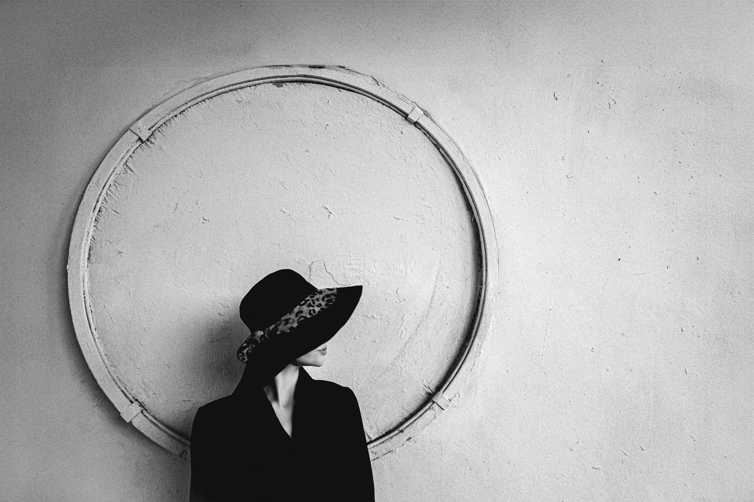 Женщина в чёрной шляпе с леопардовой лентой у круглого рельефа на стене, чёрно-белая минималистичная арт-фотография