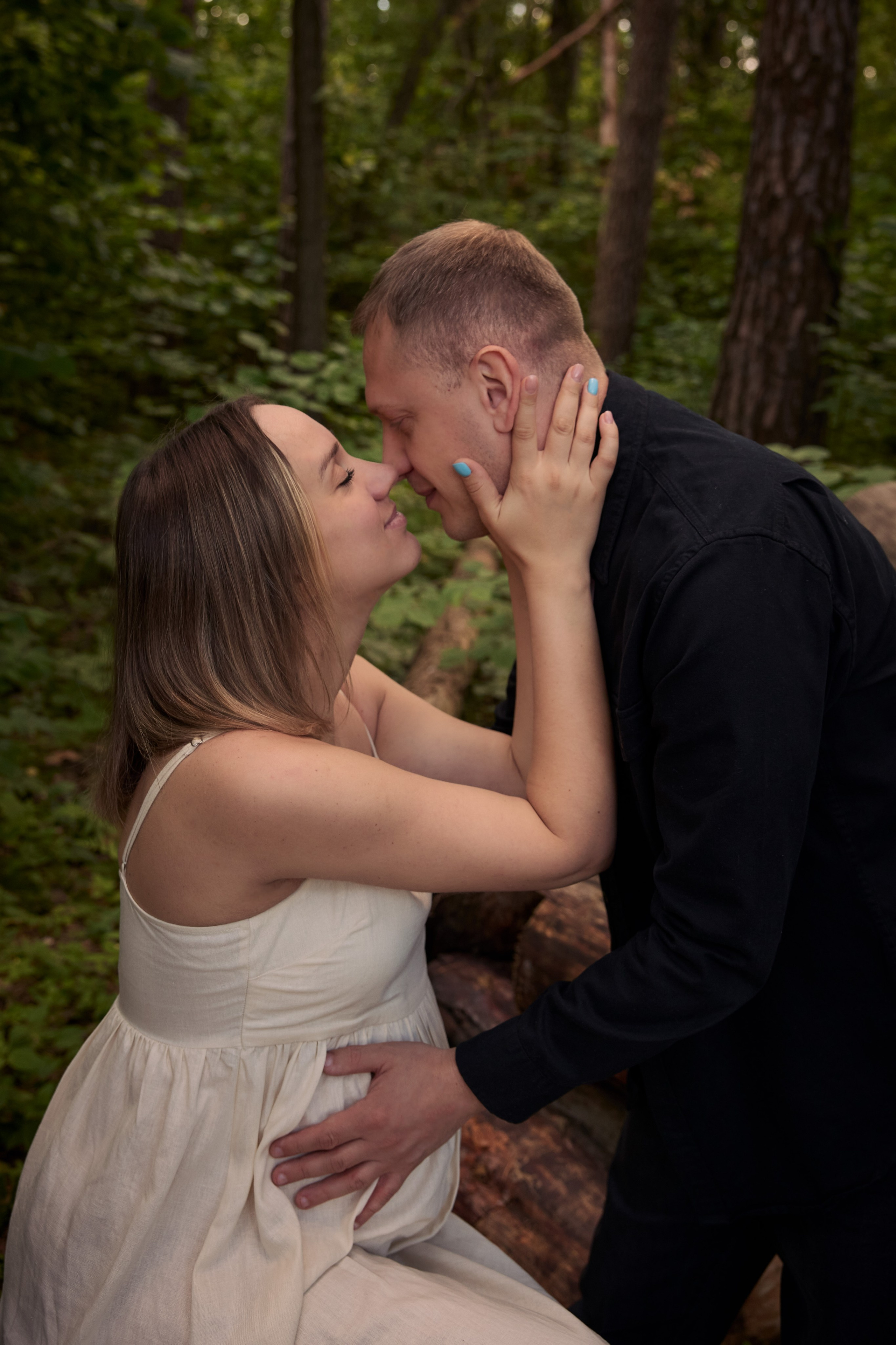 Романтический портрет будущей пары в пышном зеленом лесу: рука мужчины лежит на животе женщины, а она нежно обнимает его лицо.