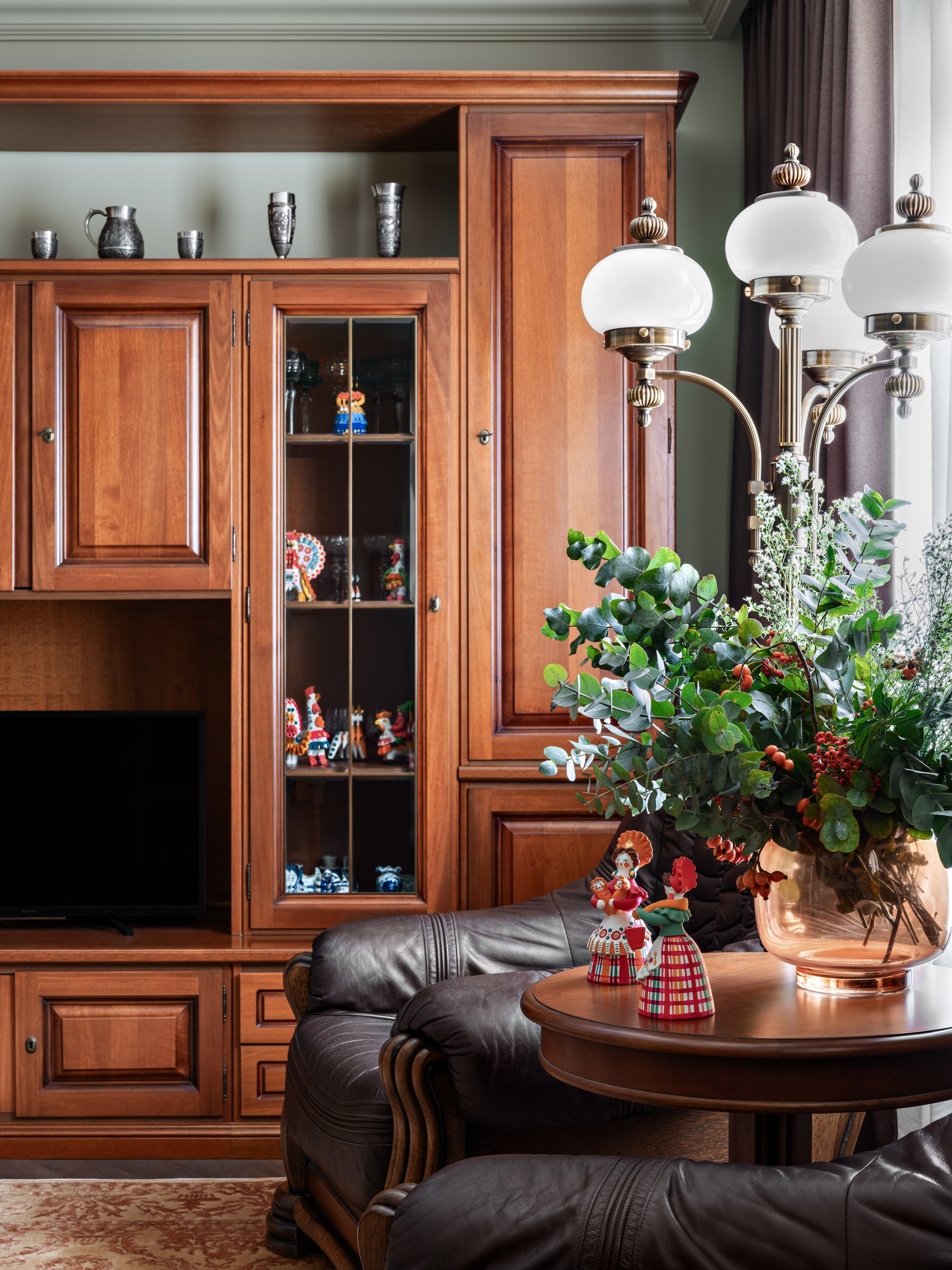 Уютная зона гостиной с деревянным шкафом-витриной, телевизором, столиком с цветочной композицией и классическим торшером; тёплый стиль интерьера подчеркивает внимание к деталям. Дизайнеры Юлия Капункина и Альбина Терещенко, стилист Алина Нелень, фотограф Сергей Денисенко.