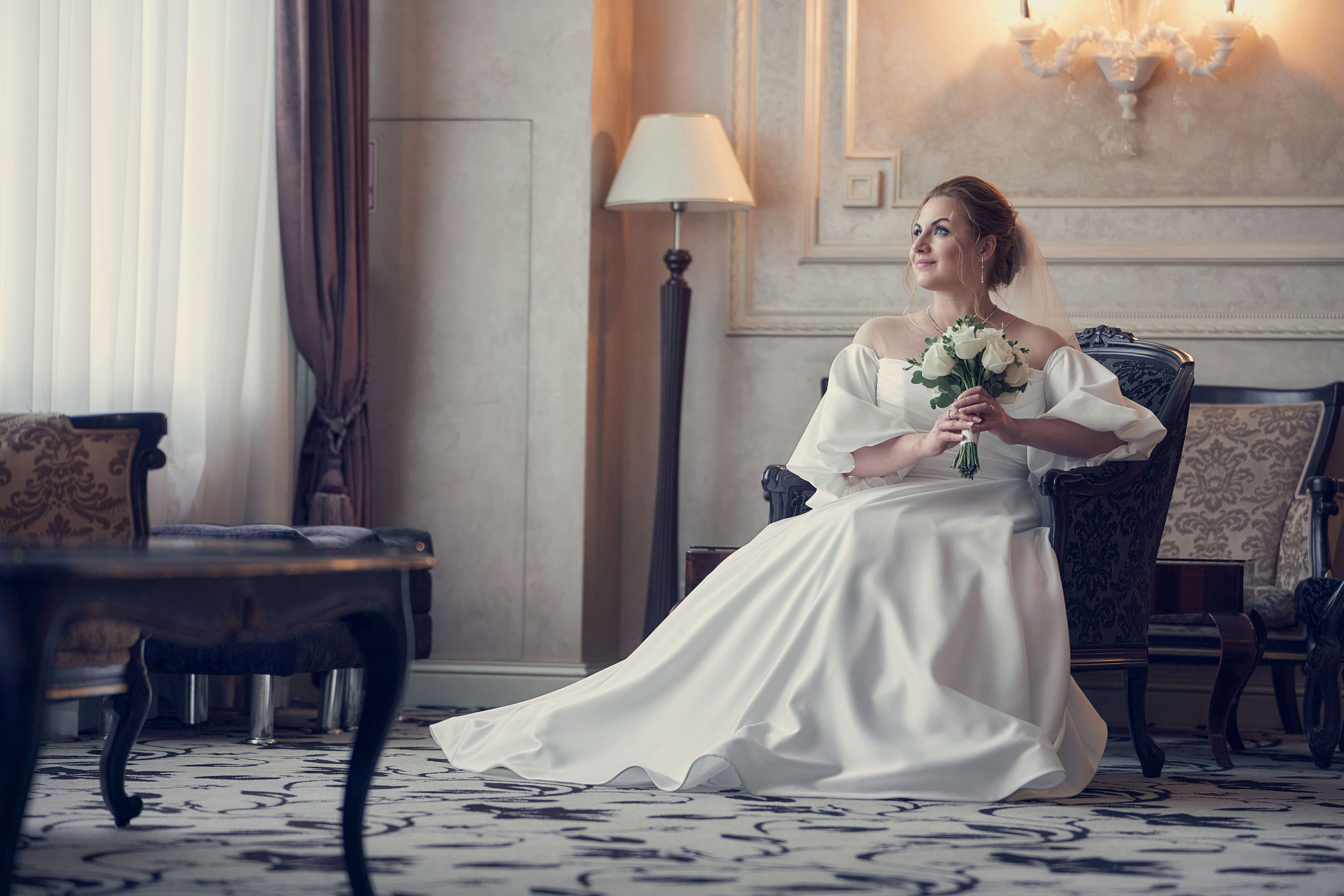 Невеста в пышном свадебном платье сидит в кресле у окна в лобби отеля