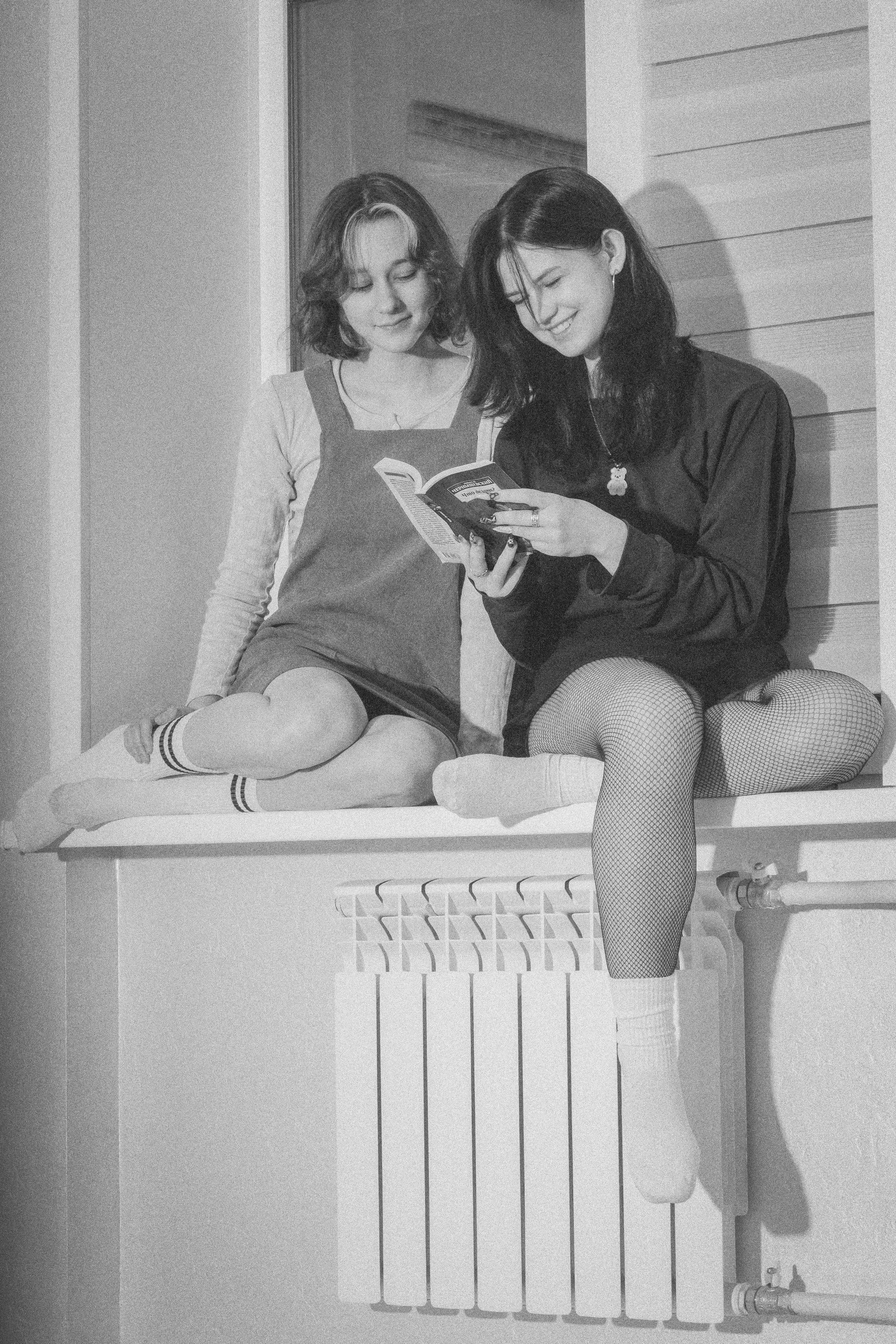 Две девушки смеются, сидя на подоконнике, держат книгу в руках — драгоценное воспоминание о сестре.