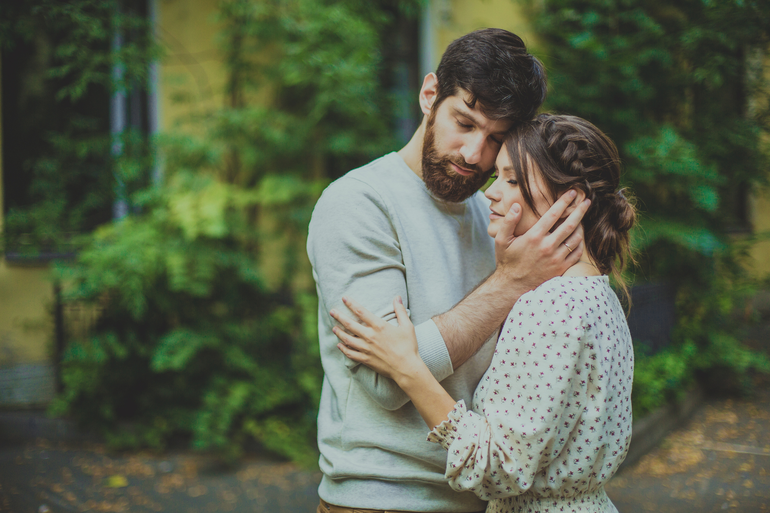 бородатый юноша нежно обнимает любимую за шею на фоне увитого зеленью дома