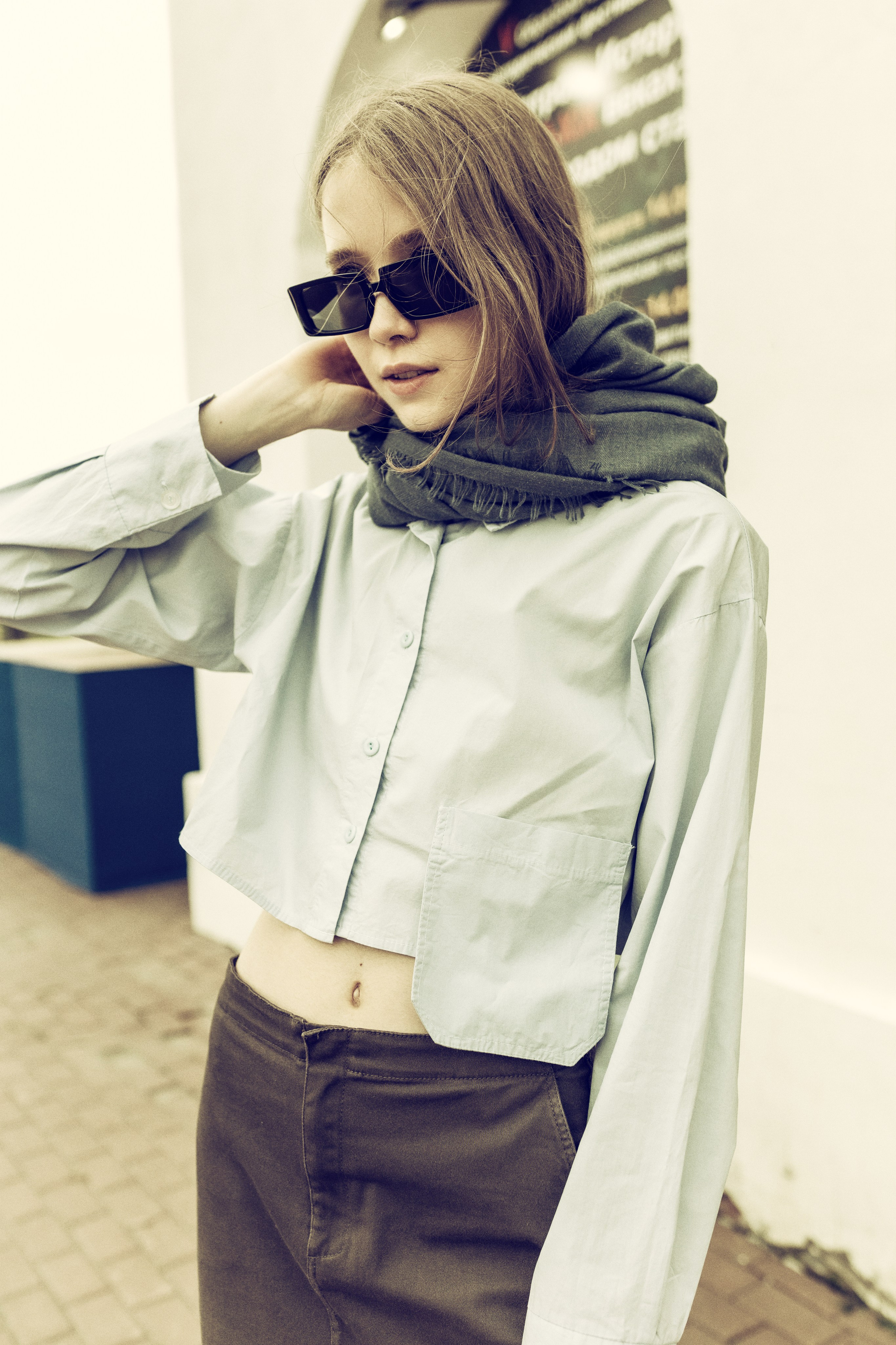 Женщина с длинными волосами, в солнцезащитных очках, сером шарфе, светлой укороченной рубашке и темных брюках, стоит на выложенной кирпичом дорожке с непринужденным, задумчивым выражением лица.