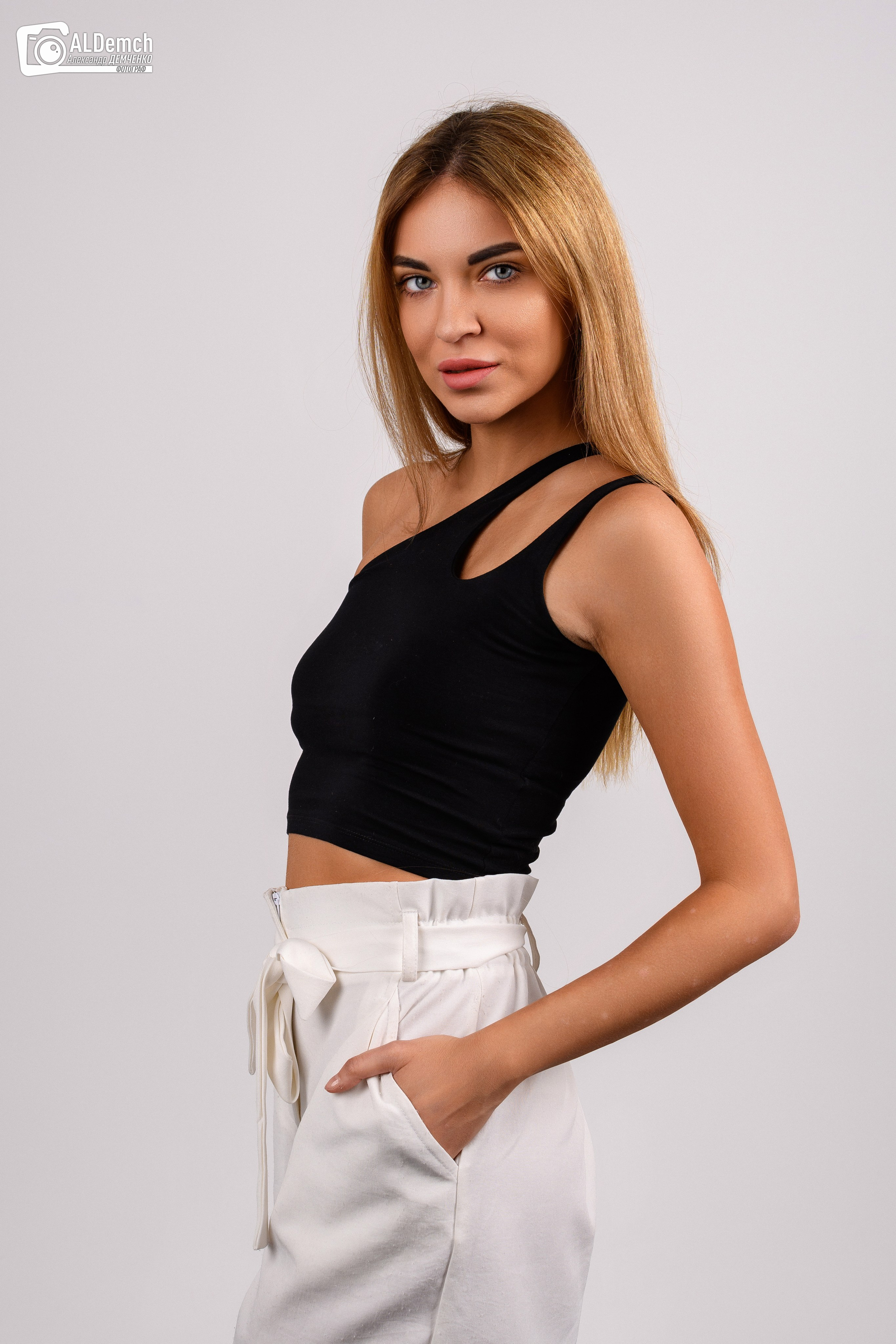 молодая женщина с длинными светлыми волосами. Она одета в черный топ с асимметричными бретелями и белые брюки с высокой талией и поясом. Женщина стоит в профиль, положив одну руку в карман брюк. Фон фотографии нейтральный, светло-серый. 