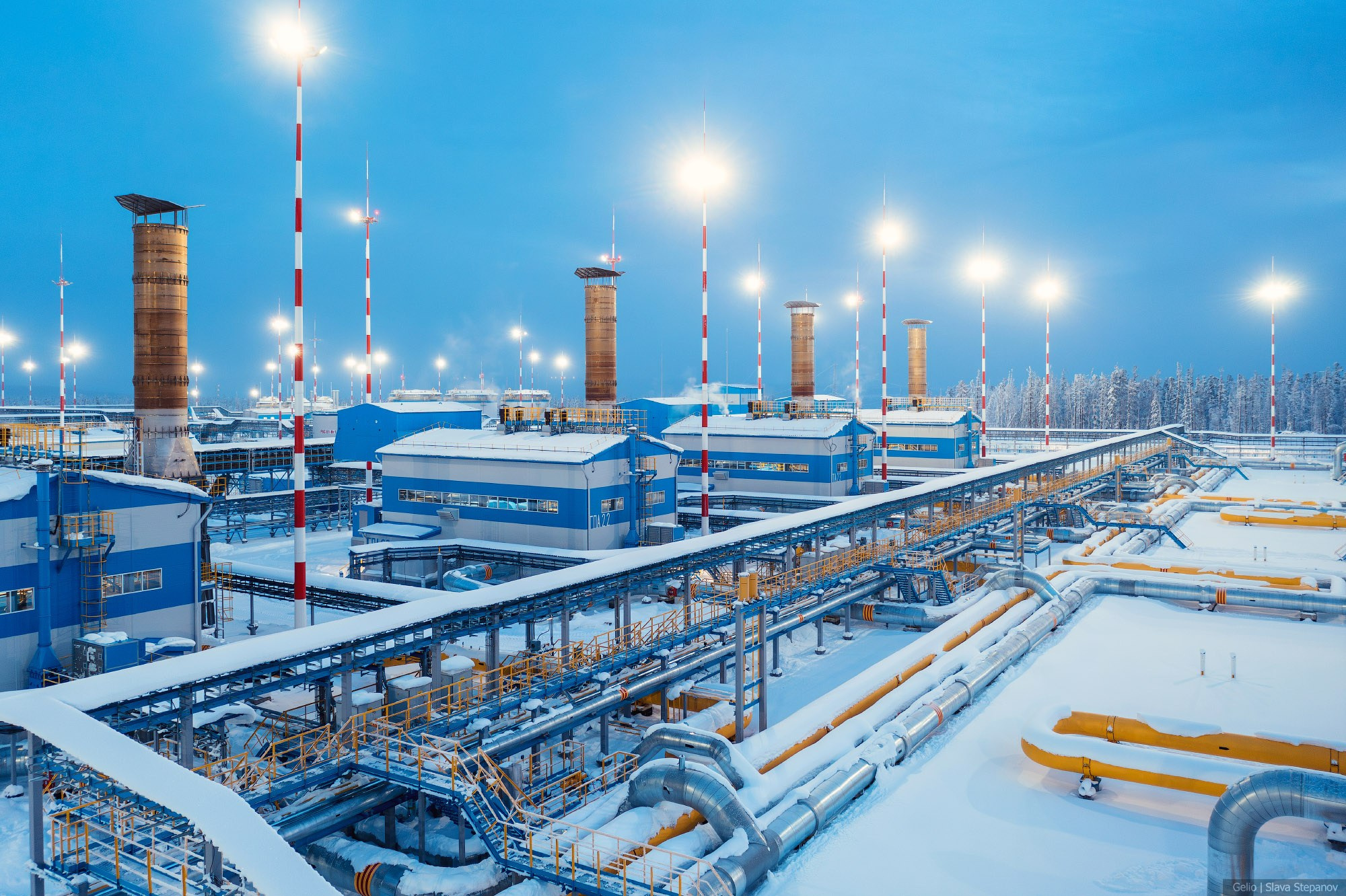 Ковыктинское газоконденсатное месторождение в зимних сумерках, заснеженная инфраструктура с высокими трубами и ярким освещением, Gelio.