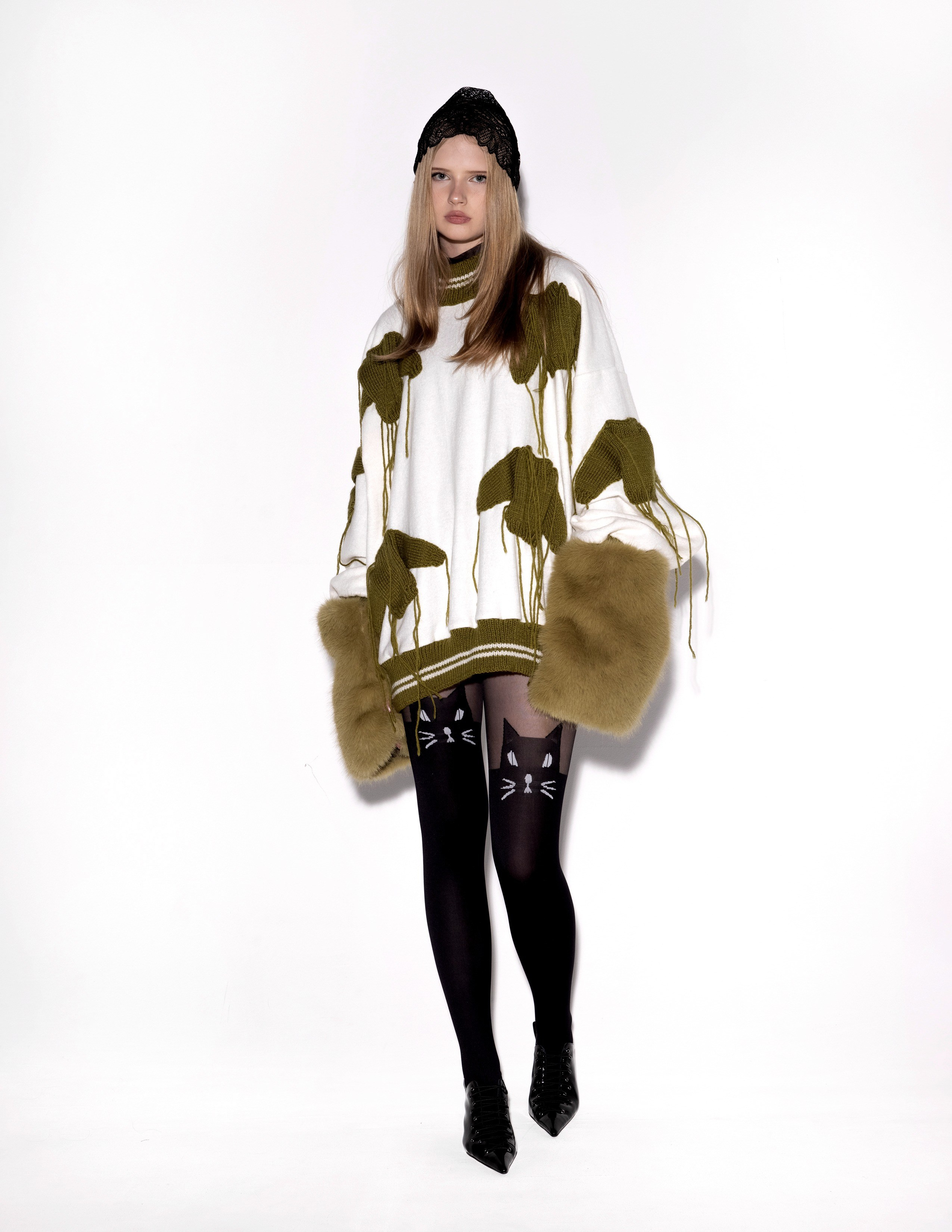 Студийная Fashion-съёмка для лукбука бренда одежды.  Ключевые детали: объемный белый свитер крупной вязки с зелеными патчами, черные колготки, глянцевые черные ботинки и пушистые меховые манжеты оливкового цвета . Образ дополнен черной шапкой с кружевной отделкой. 