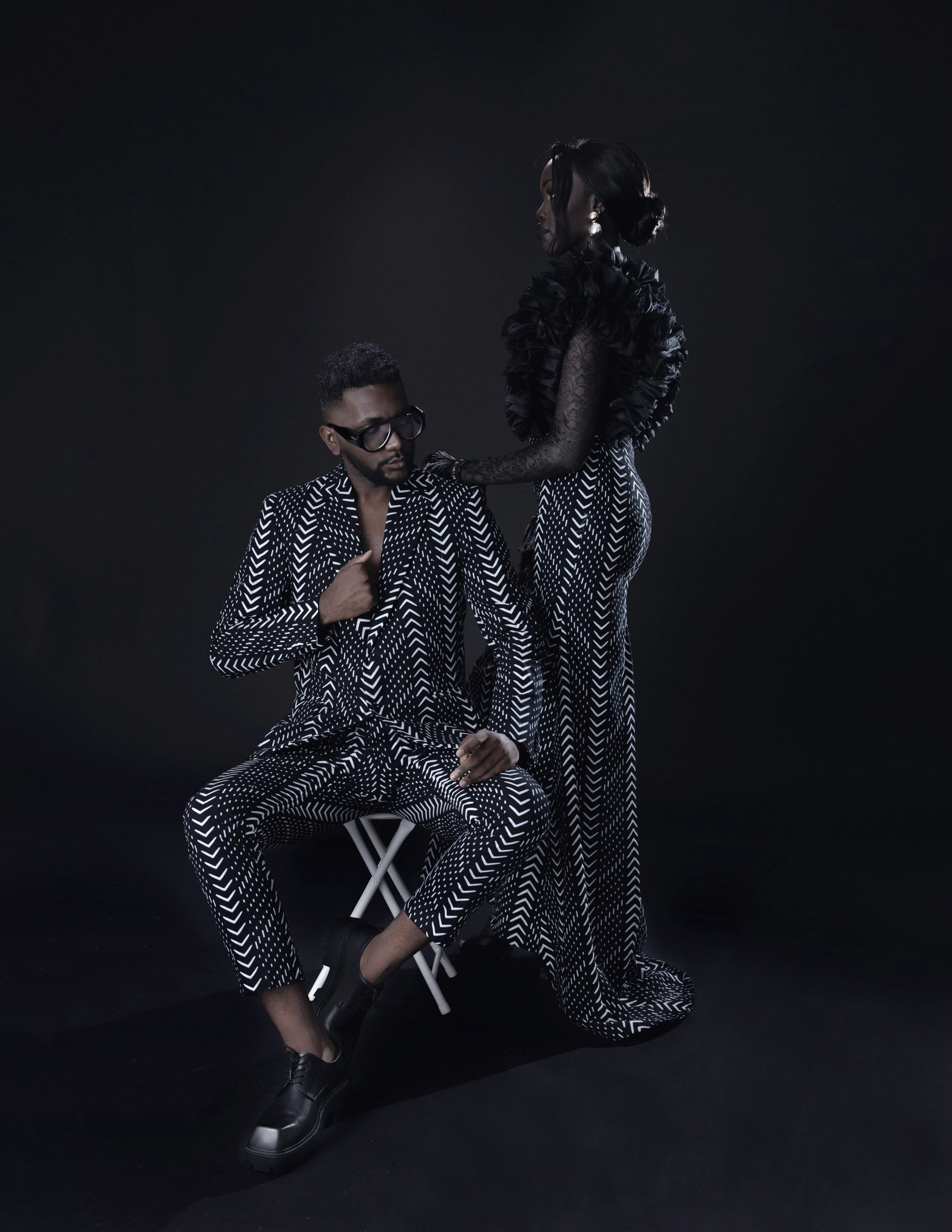 Студийная фешн-съёмка для известного модельера одежды haute couture . Темнокожие мужчина и девушка  на черном фоне в эффектных одеждах с одинаковым геометрическим черно-белым узором «елочка». Мужчина в строгом двубортном костюме, массивных очках и грубых ботинках сидит на белом складном стуле. Девушка в длинном платье в тон с объемными черными рукавами стоит позади него, положив руку ему на плечо. Фотография передает элегантность и драматизм, характерные для высокой моды. Съёмка фешн-фотографа Станислава Ефимова в ЮАР.