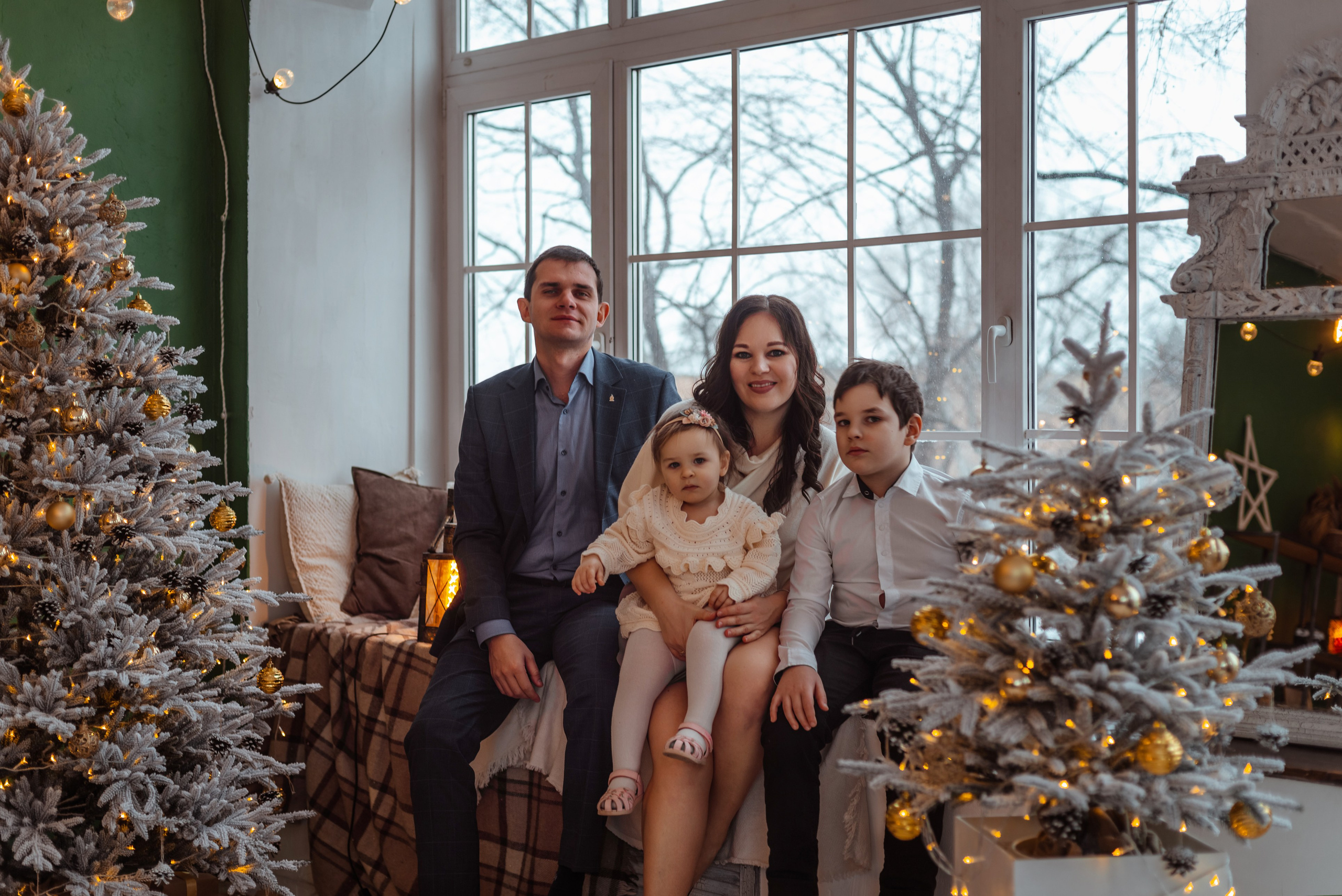 Семья из четырех человек позирует на фоне новогодних елок и украшенного окна.
