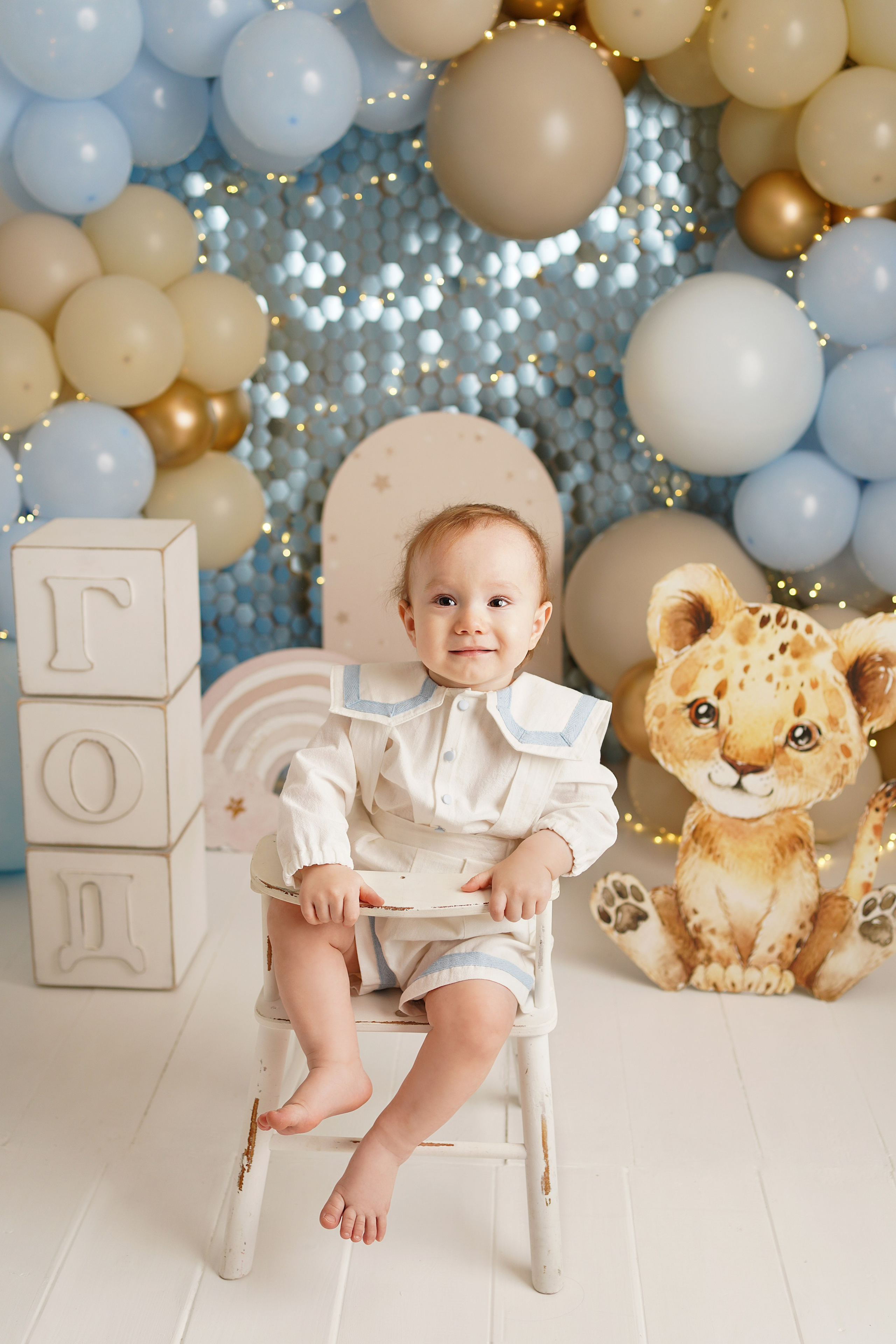 На фото малыш на фотосессии годик. На нем красивый наряд, вокруг много игрушек и воздушных шариков.