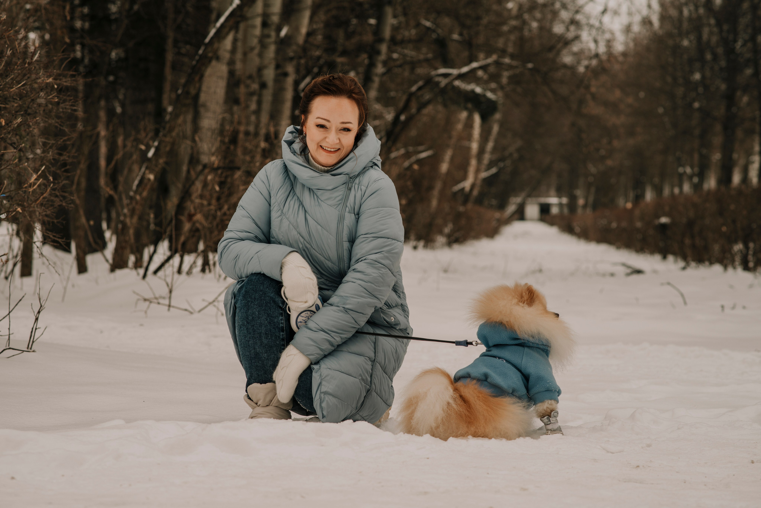 Девушка с рыжими волосами позирует на фоне зимнего леса в голубом пуховике