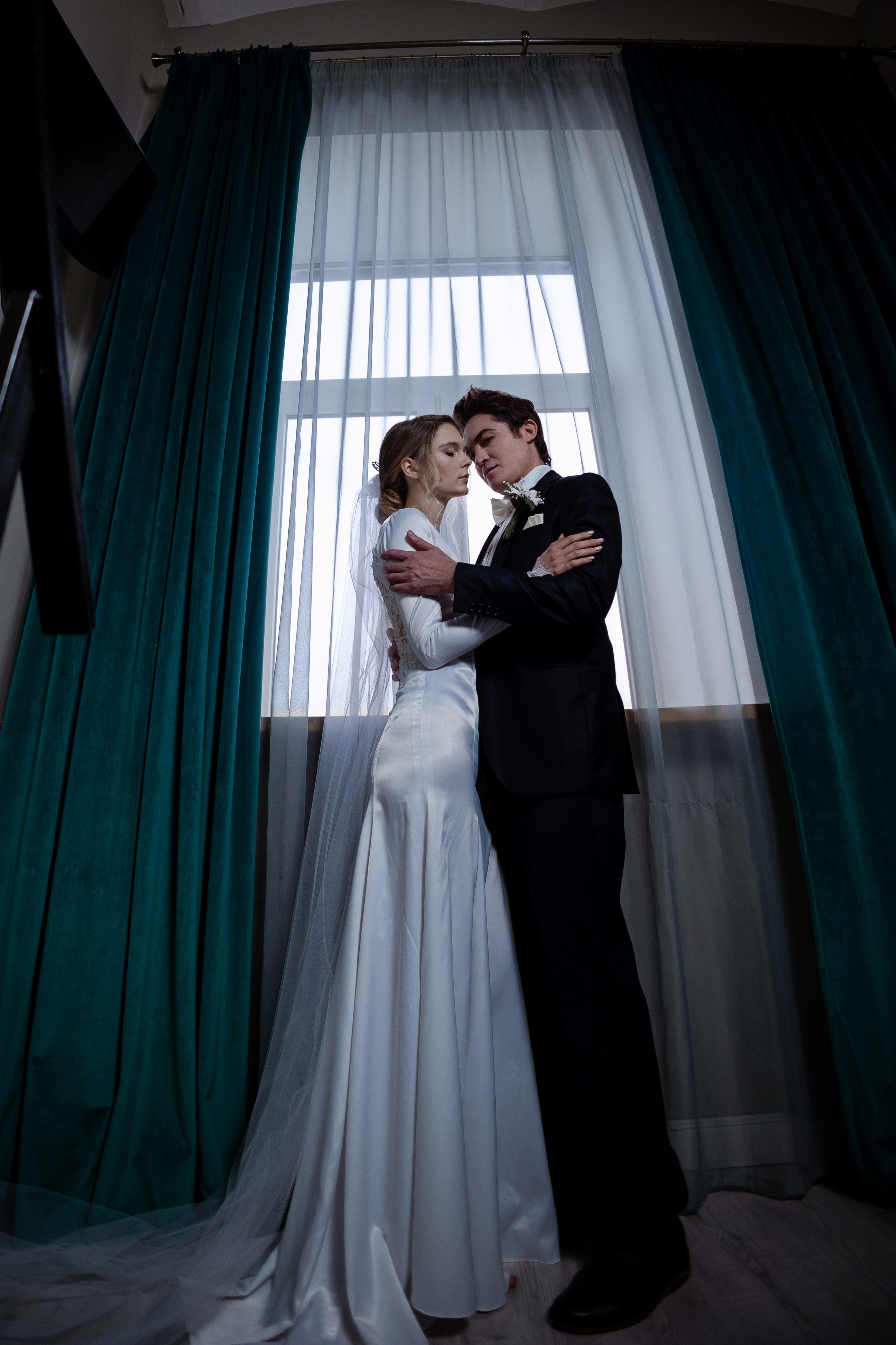 Жених и невеста обнимают друг друга на фоне окна и изумрудных штор.