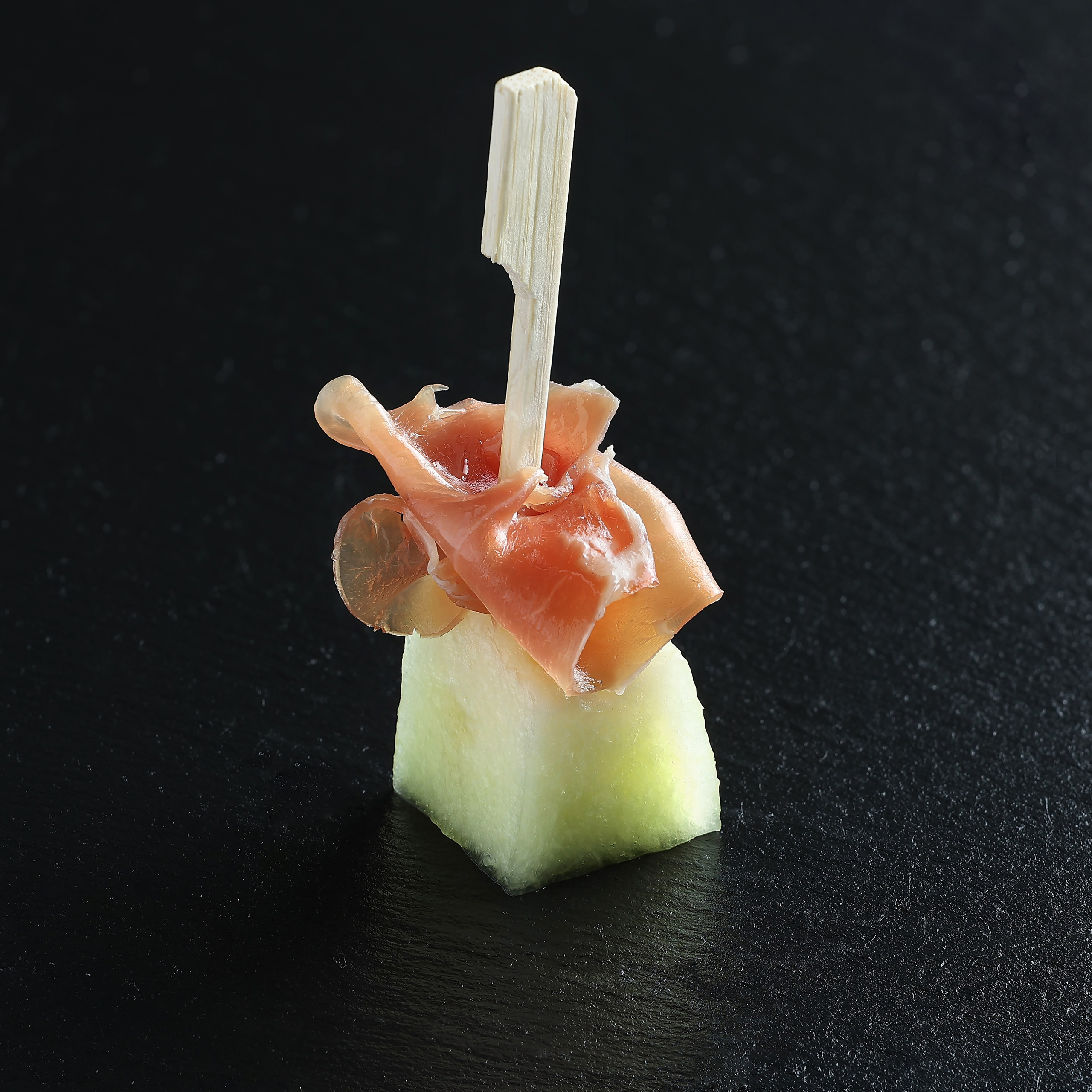 Закуска из дыни и пармской ветчины, скреплённая деревянной шпажкой, на тёмном фоне.