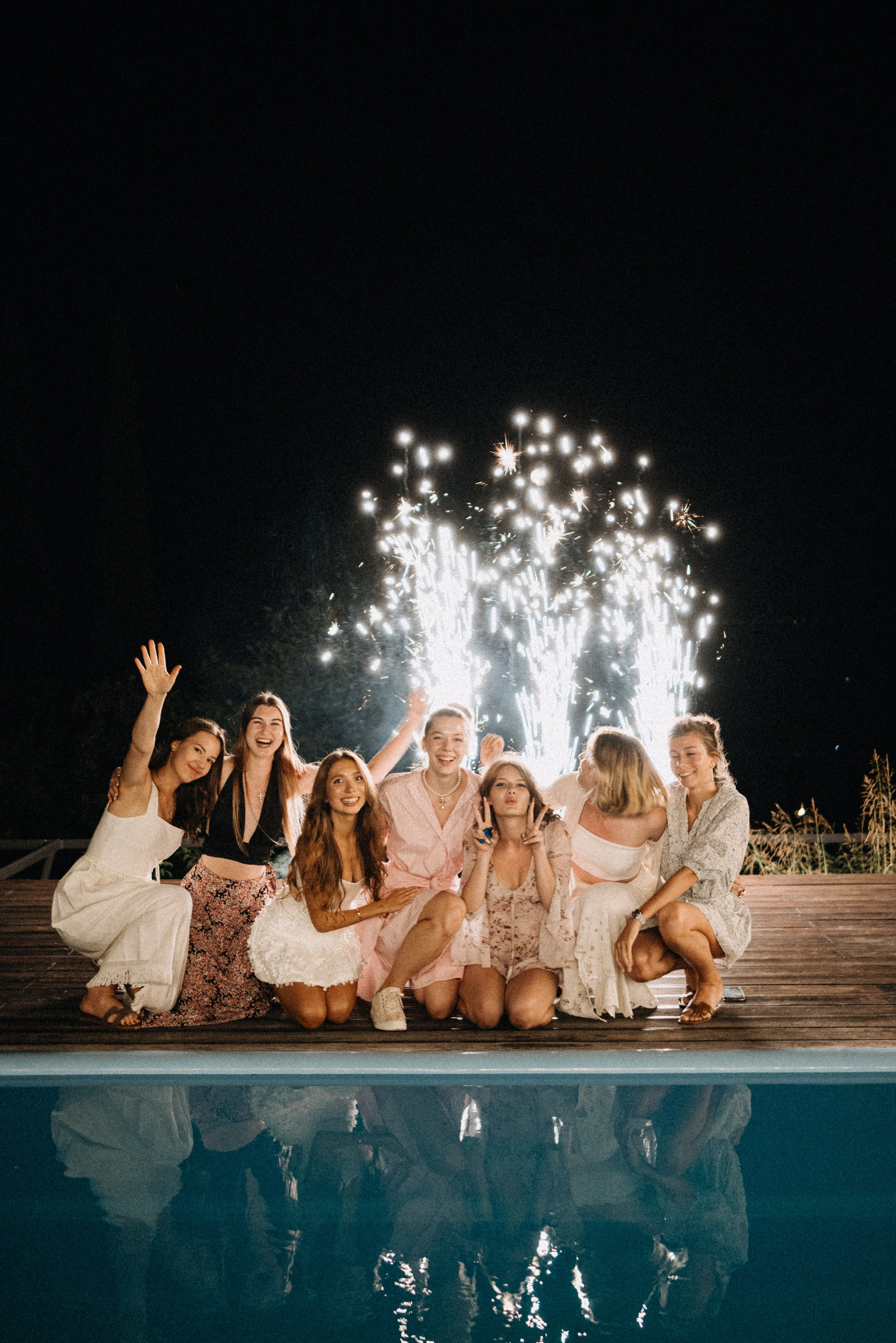 Фотография семерых празднично одетых девушек на фоне горящего фейерверка, сидящих у бассейна