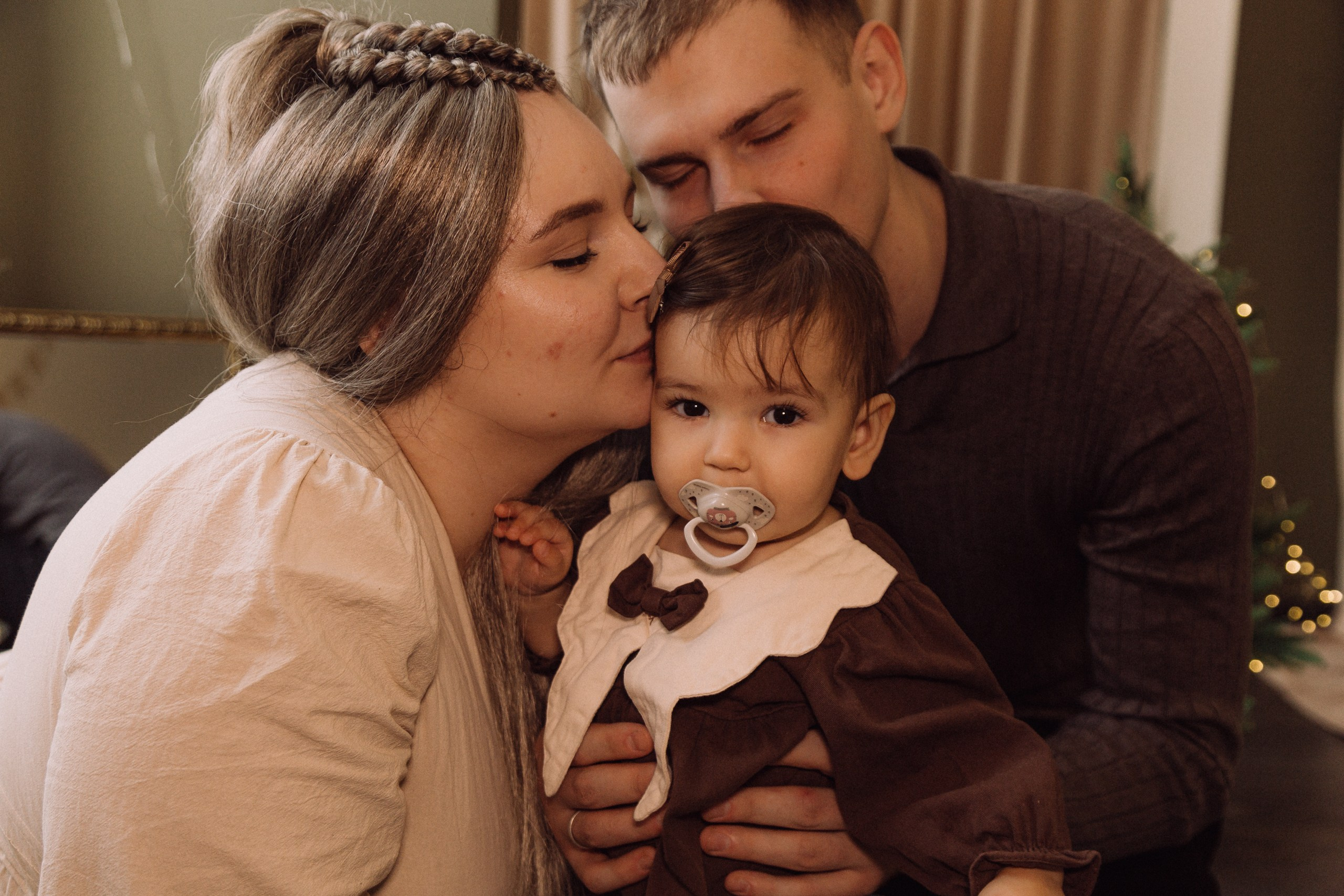 Родители держат малыша между собой, мама целует щёку ребёнка, семейный портрет в интерьере.