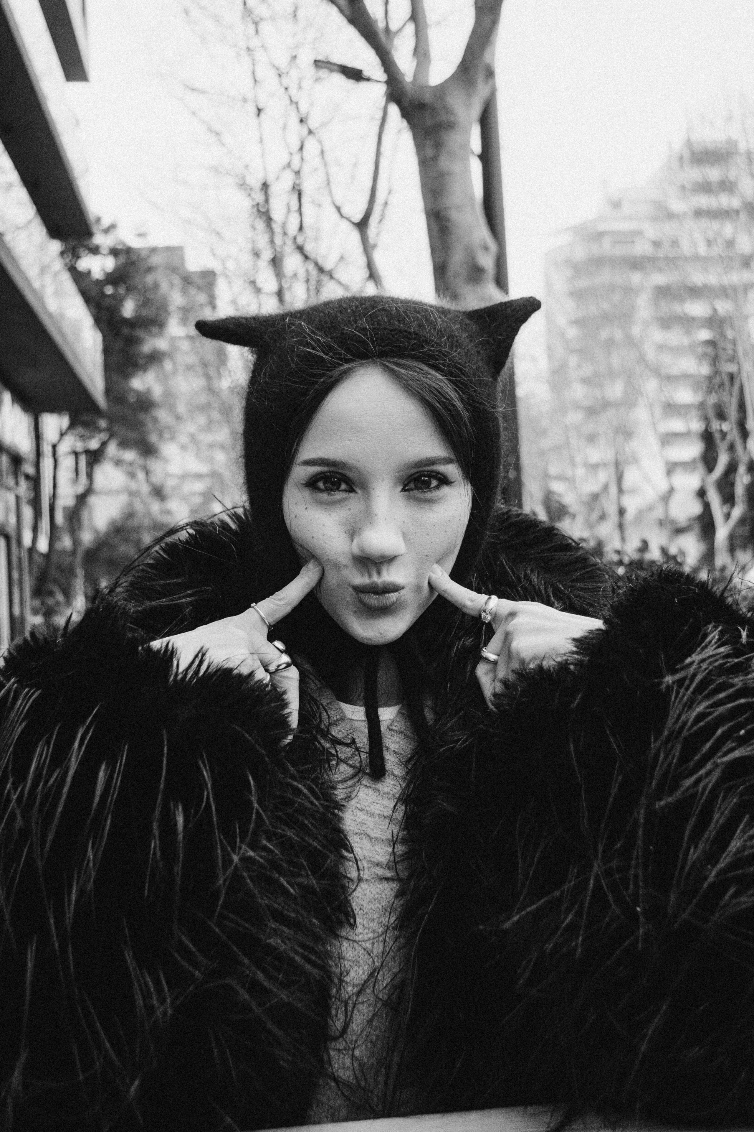 Игривый городской черно-белый портрет девушки в шубе и вязаной шапке с ушками. Девушка смотри прямо на нас и утыкается двумя указательными пальцами себе в щеки