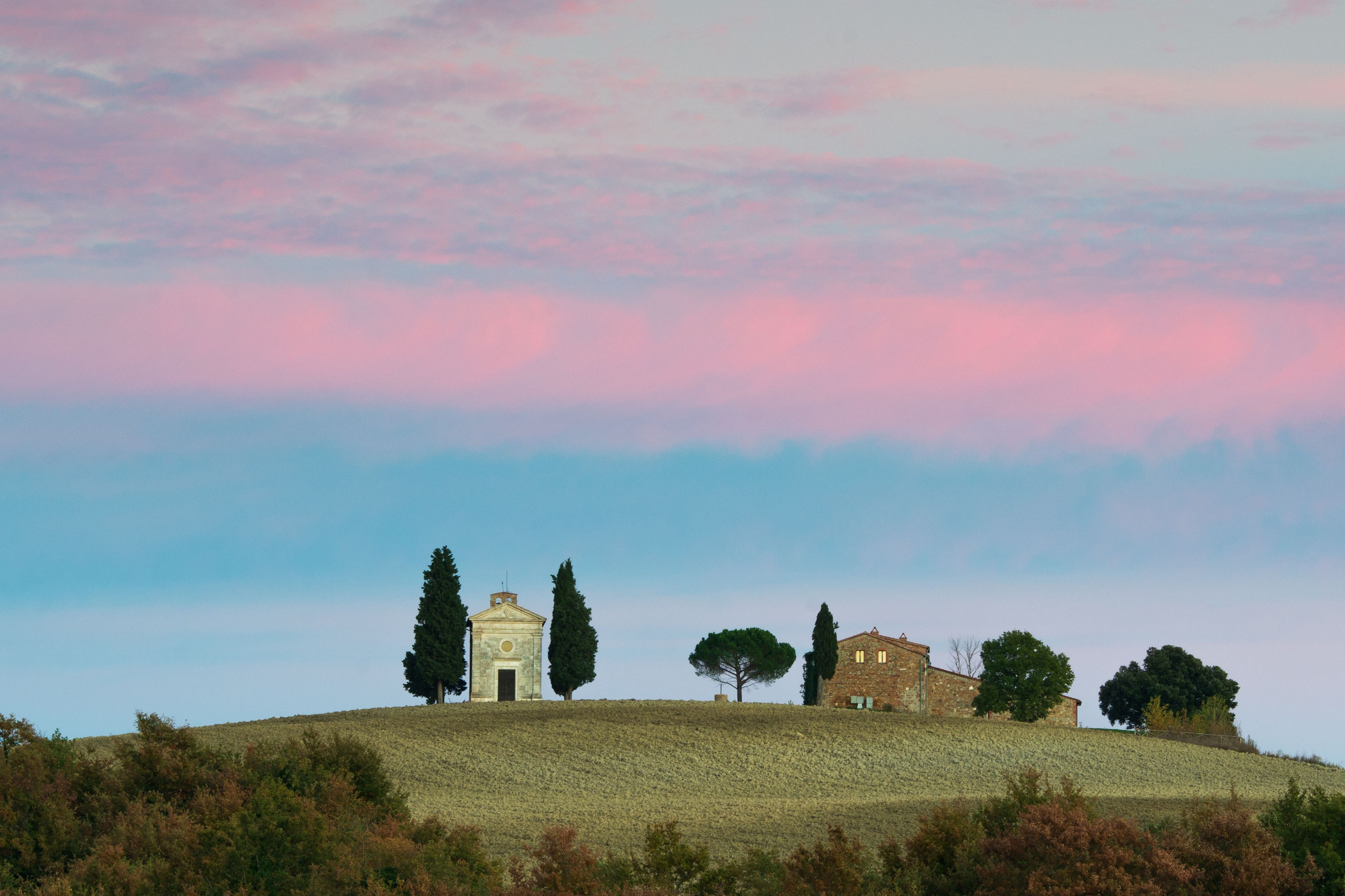 Классический пейзаж Тосканы: иконическая часовня Виталета и фермерский дом на вершине холма, окруженные кипарисами, на фоне драматичного розово-голубого неба на закате, спокойная атмосфера.
