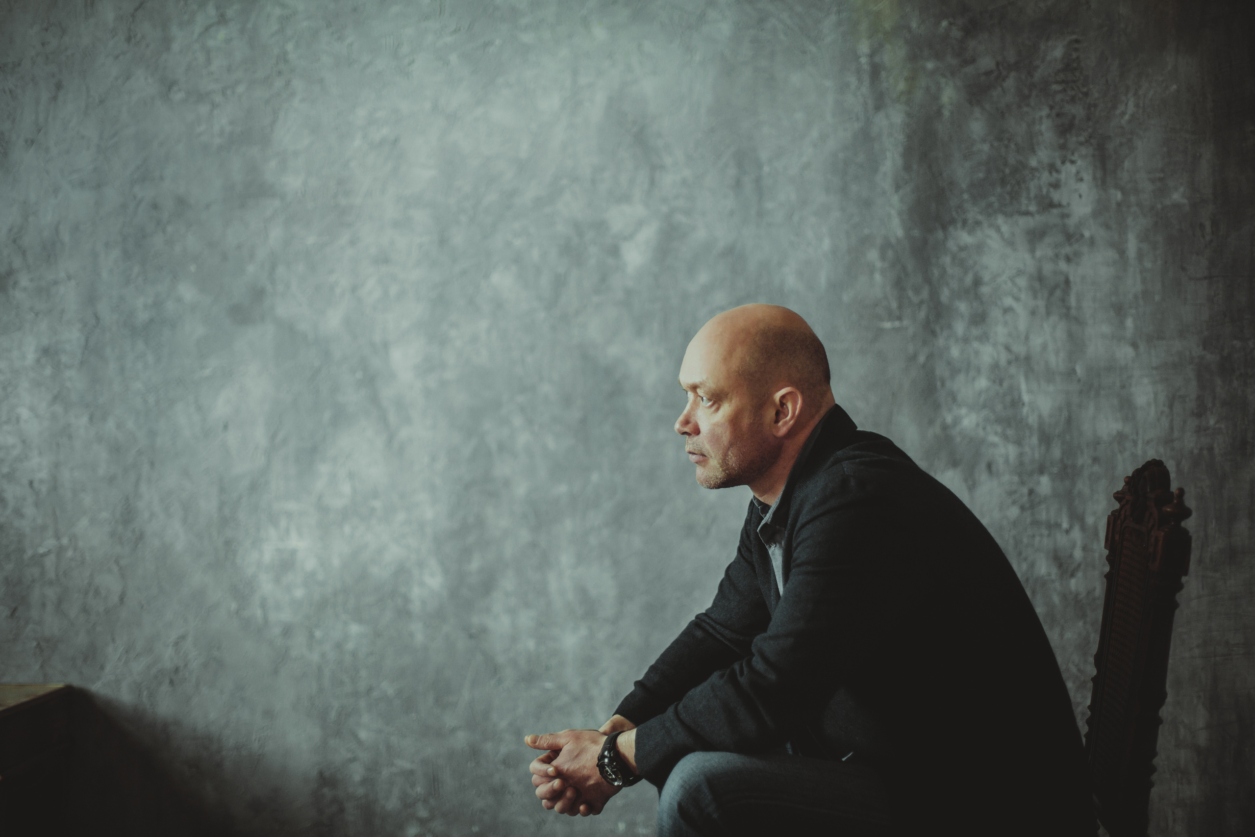 профиль лысого мужчины сидящего на стуле у серой стены