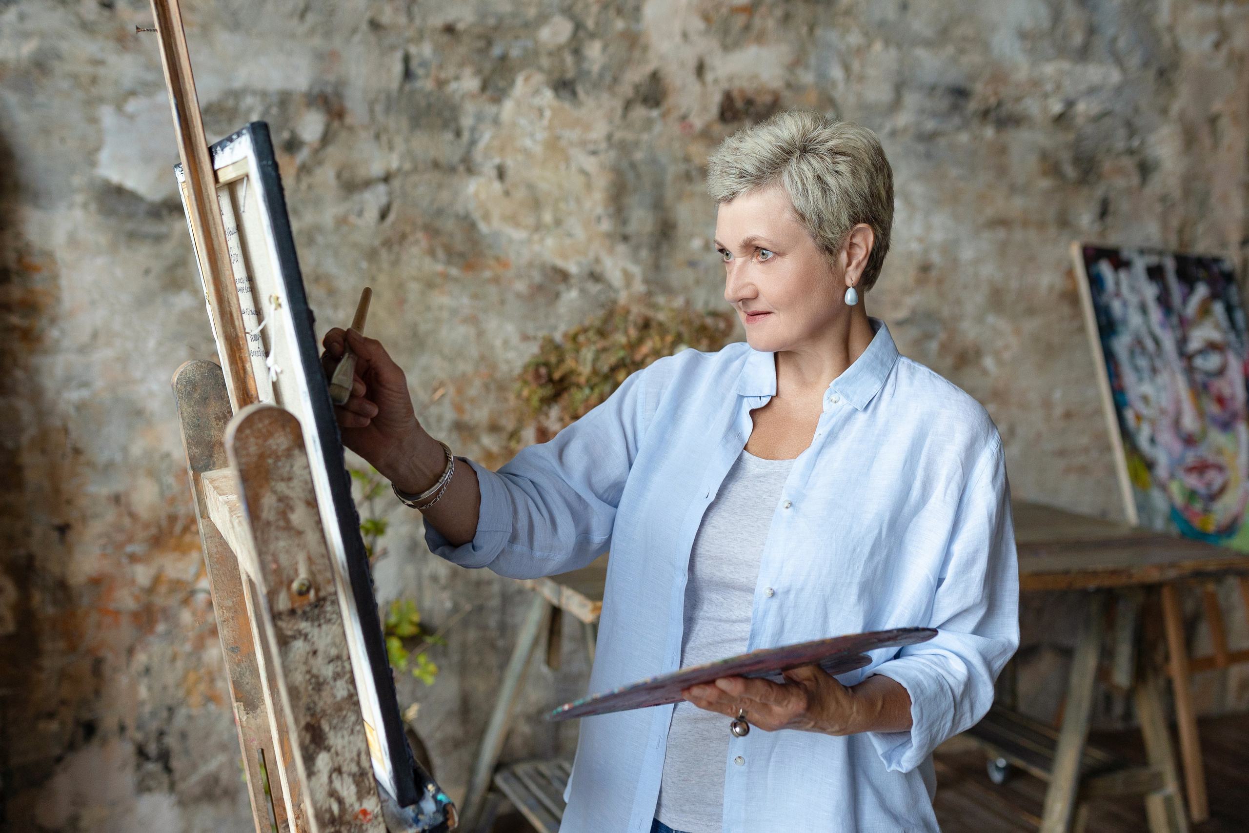 Художница стоит у пюпитра с палитрой и рисует.