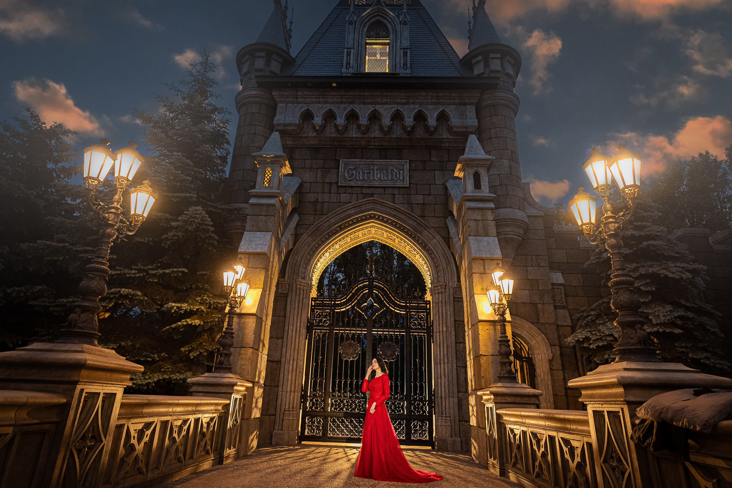 Девушка в роскошном красном платье стоит у главных ворот замка Гарибальди, подсвеченных тёплым сиянием фонарей.