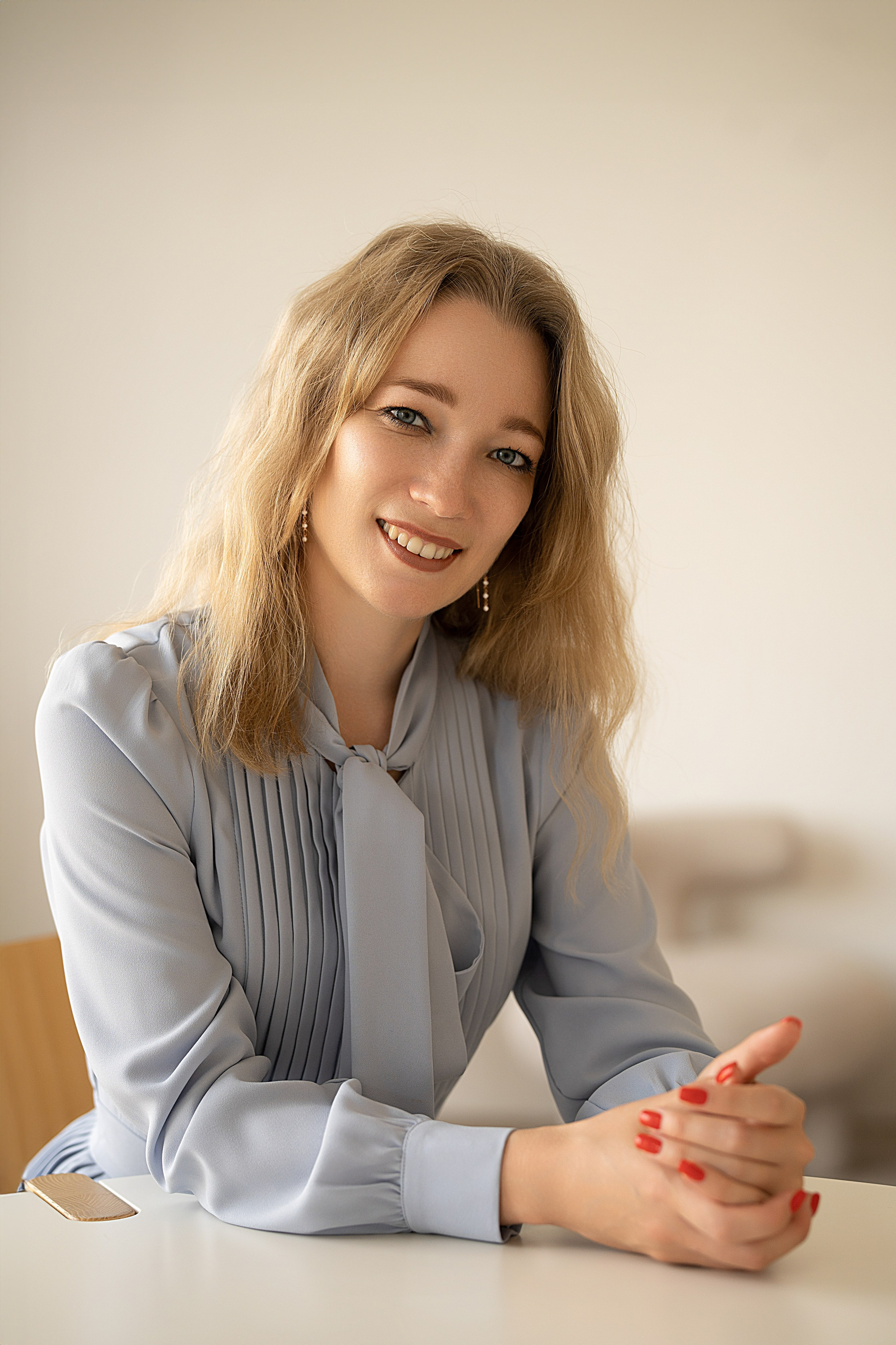 Бизнес-портрет женщины. Она сидит за столом и смотрит прямо в кадр. Женщина одета в голубое платье.