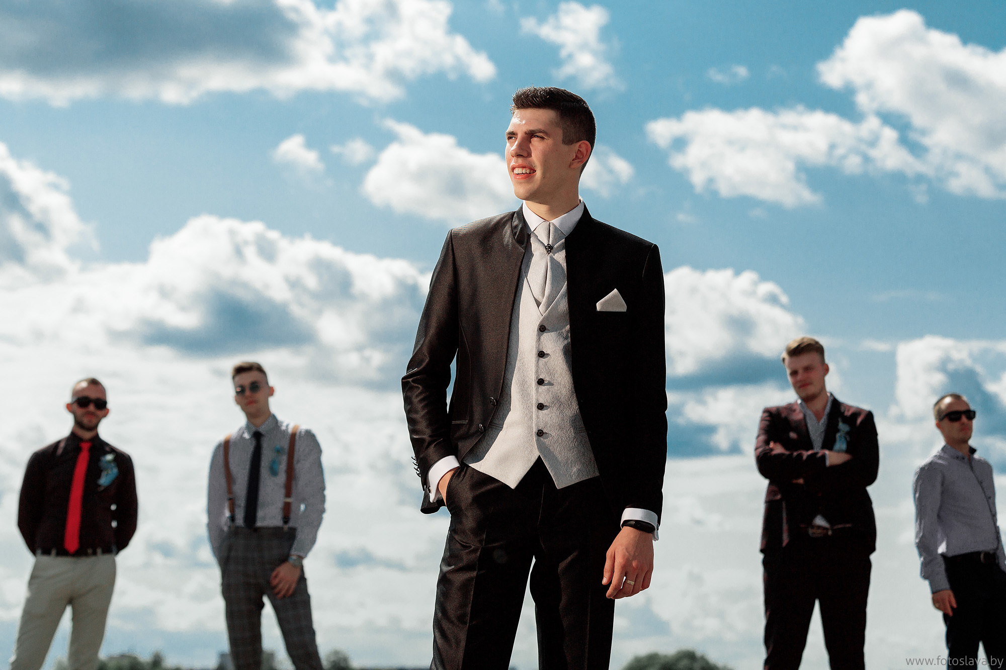 жених на фоне синего неба с облаками и друзей на заднем плане.