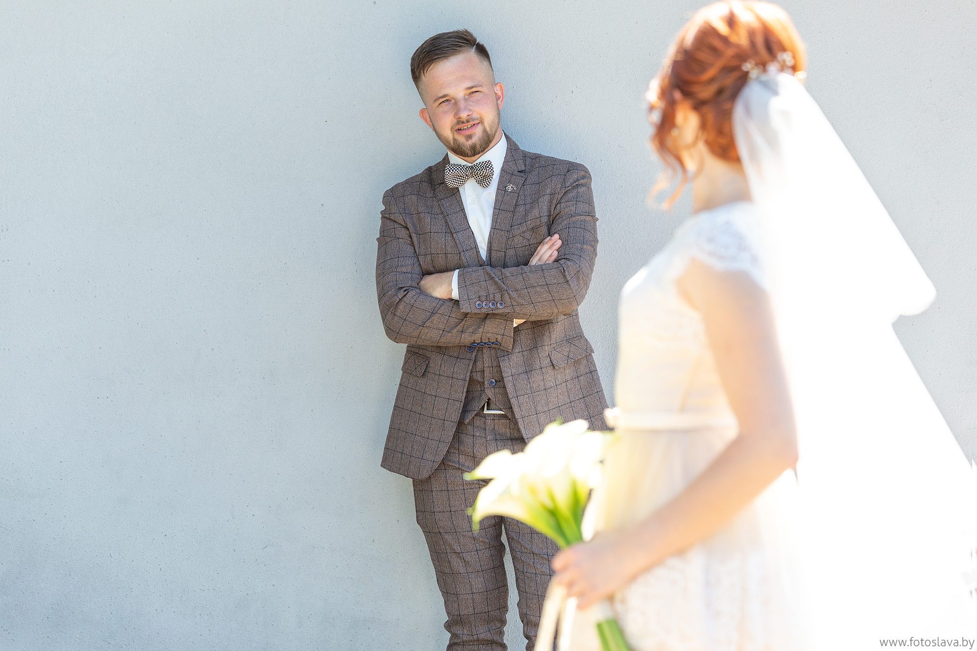 жених, прислонившись к стене на заднем плане в резкости смотрит на невесту на переднем плане в расфокусе, которая обернувшись, смотрит на него.