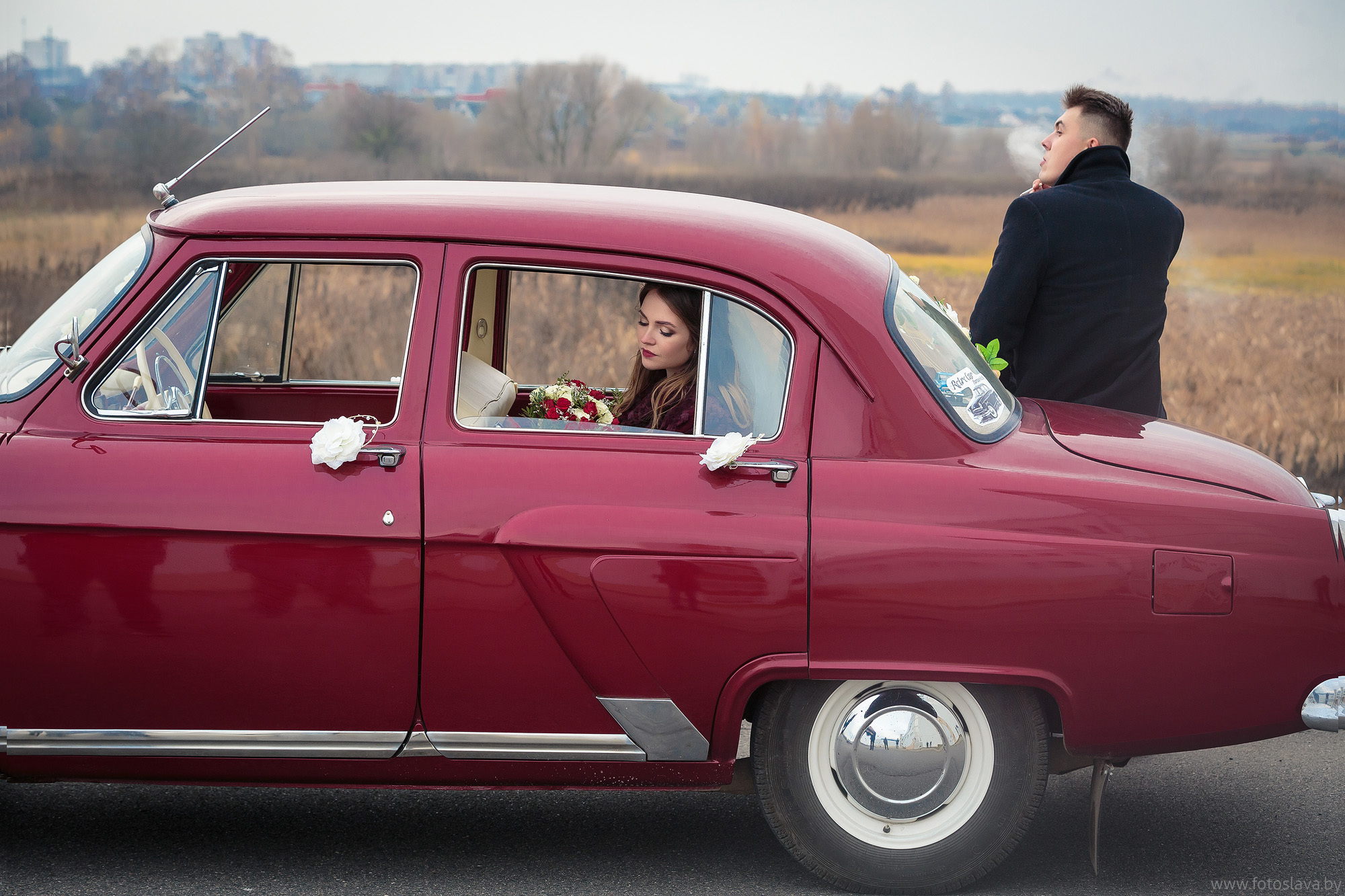 жених курит, сидя  на заднем крыле автомобиля, невеста в автомобиле.