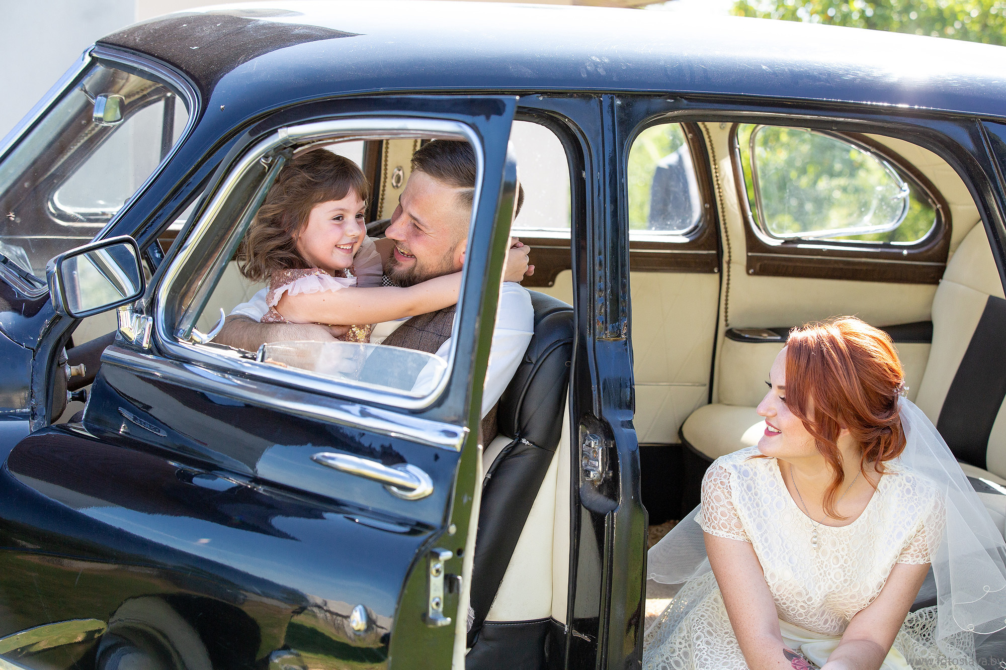 жених сидит в машине и обнимает ребенка, невеста рядом смотрит на них.