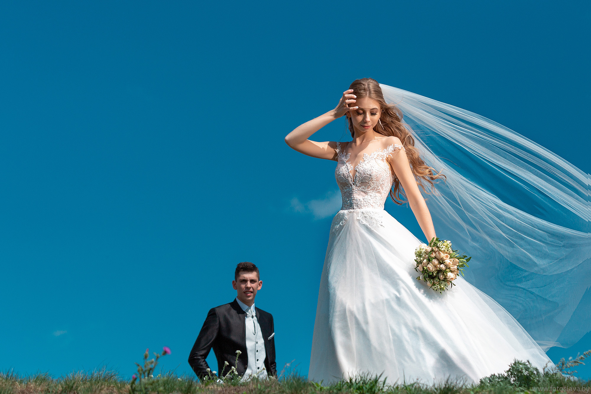 невеста в полный рост на фоне синего неба удерживает одной рукой фату, и чуть дальше на горизонте на нее смотрит жених.