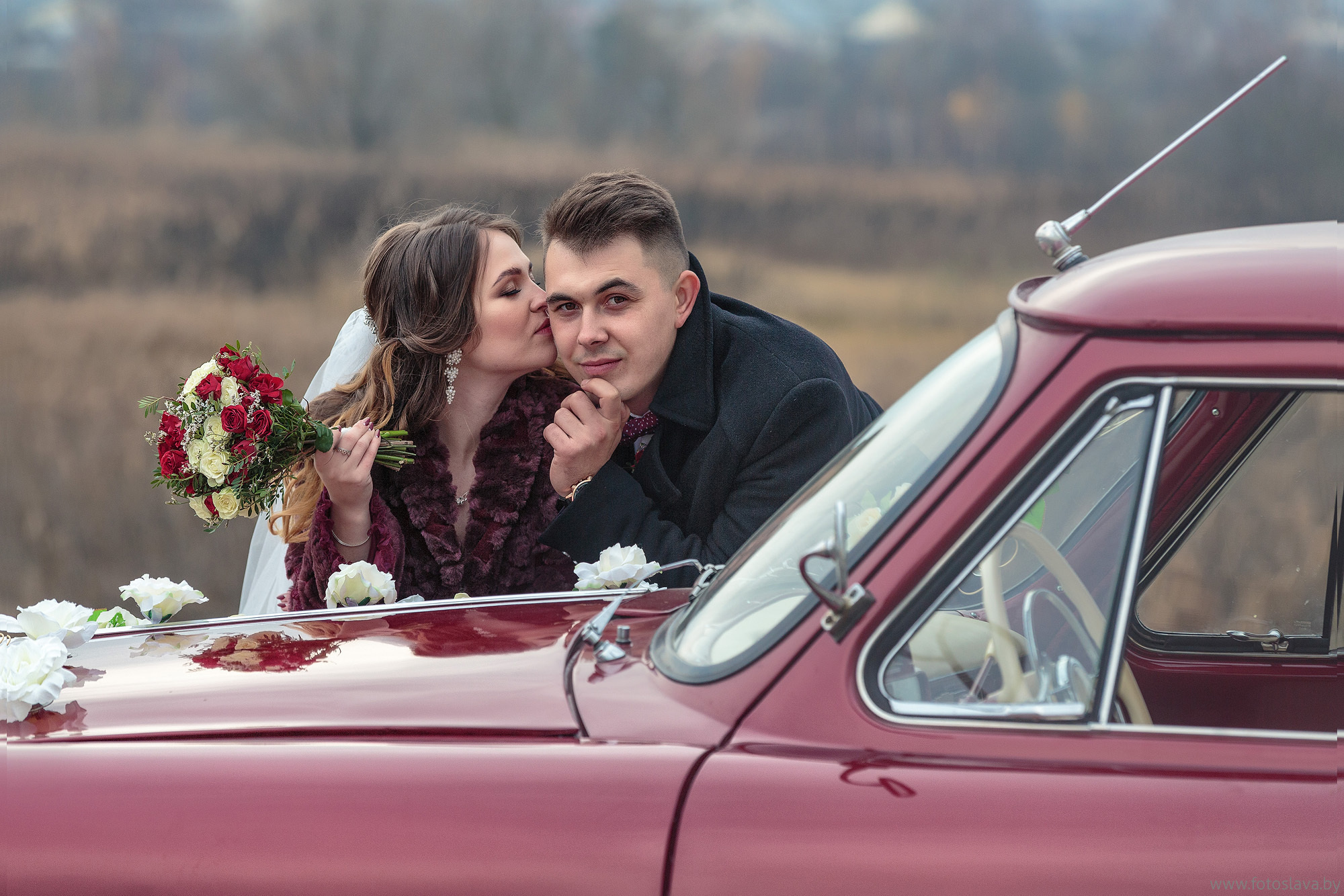 жених с невестой облокотились на капот автомобиля , невеста целует жениха в щеку.