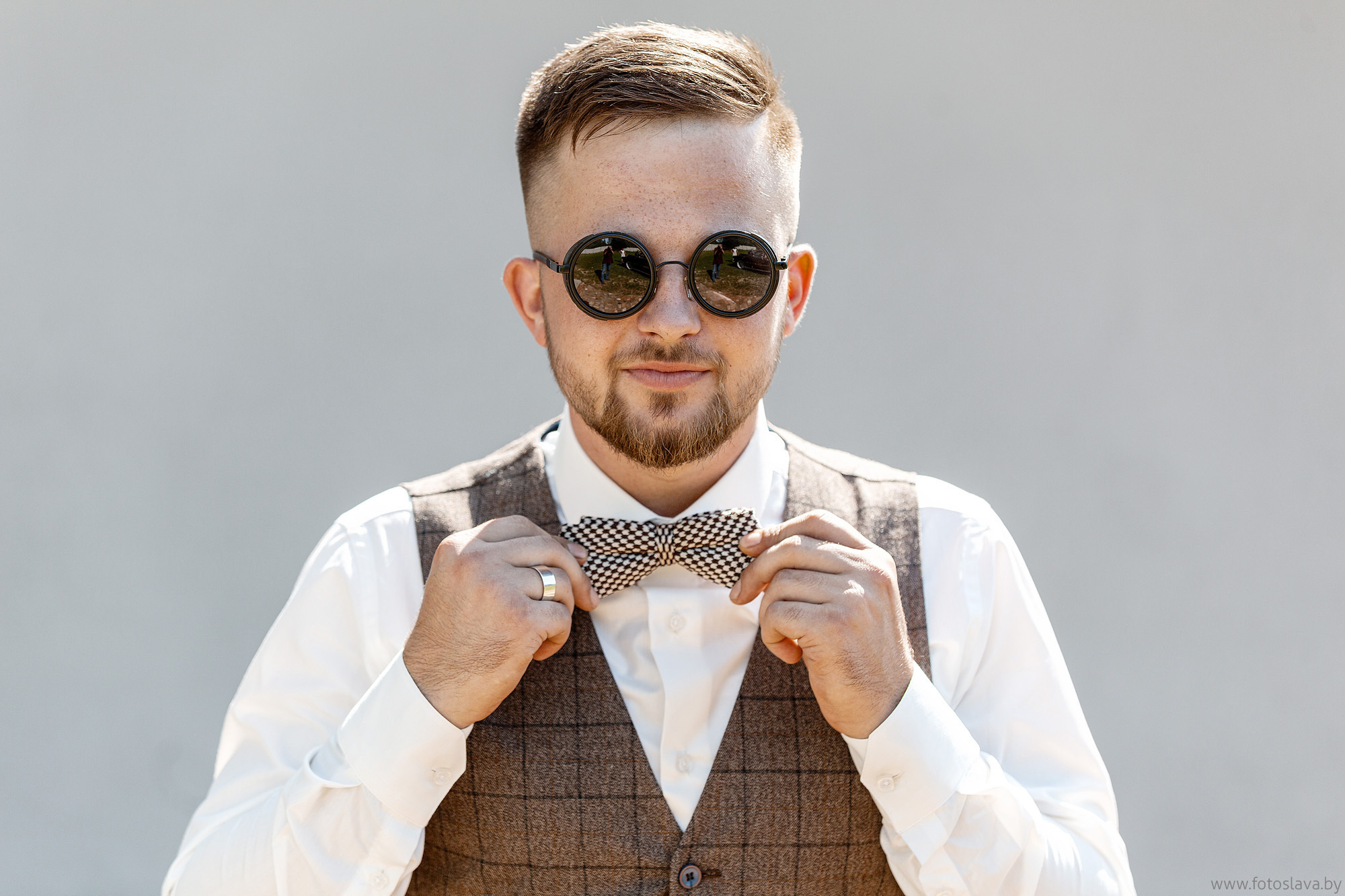 жених в жилетке и черных круглых очках с бабочкой , снят по пояс, позирует фотографу).