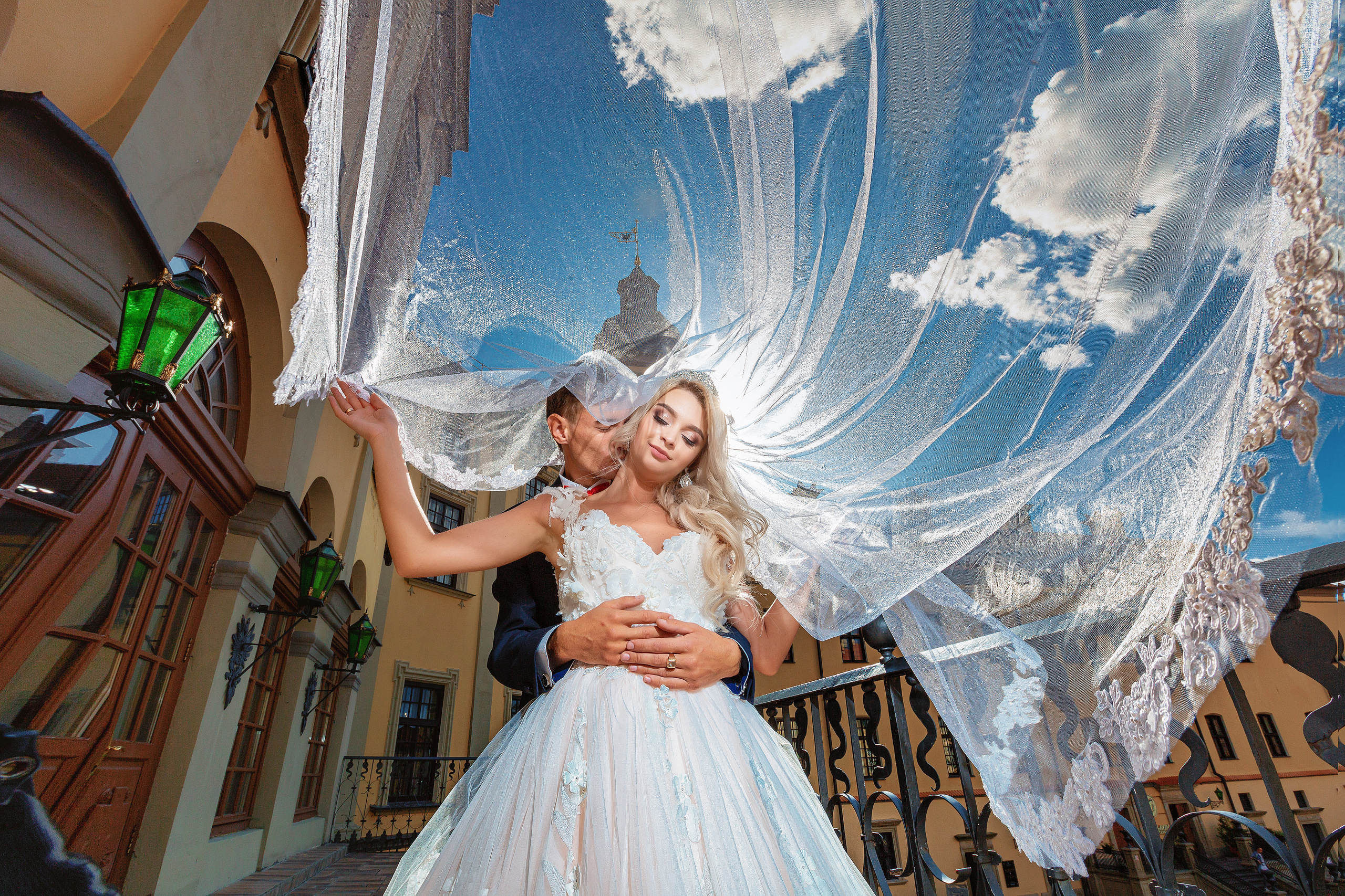 жених сзади обнимает невесту на фоне развевающейся фаты.