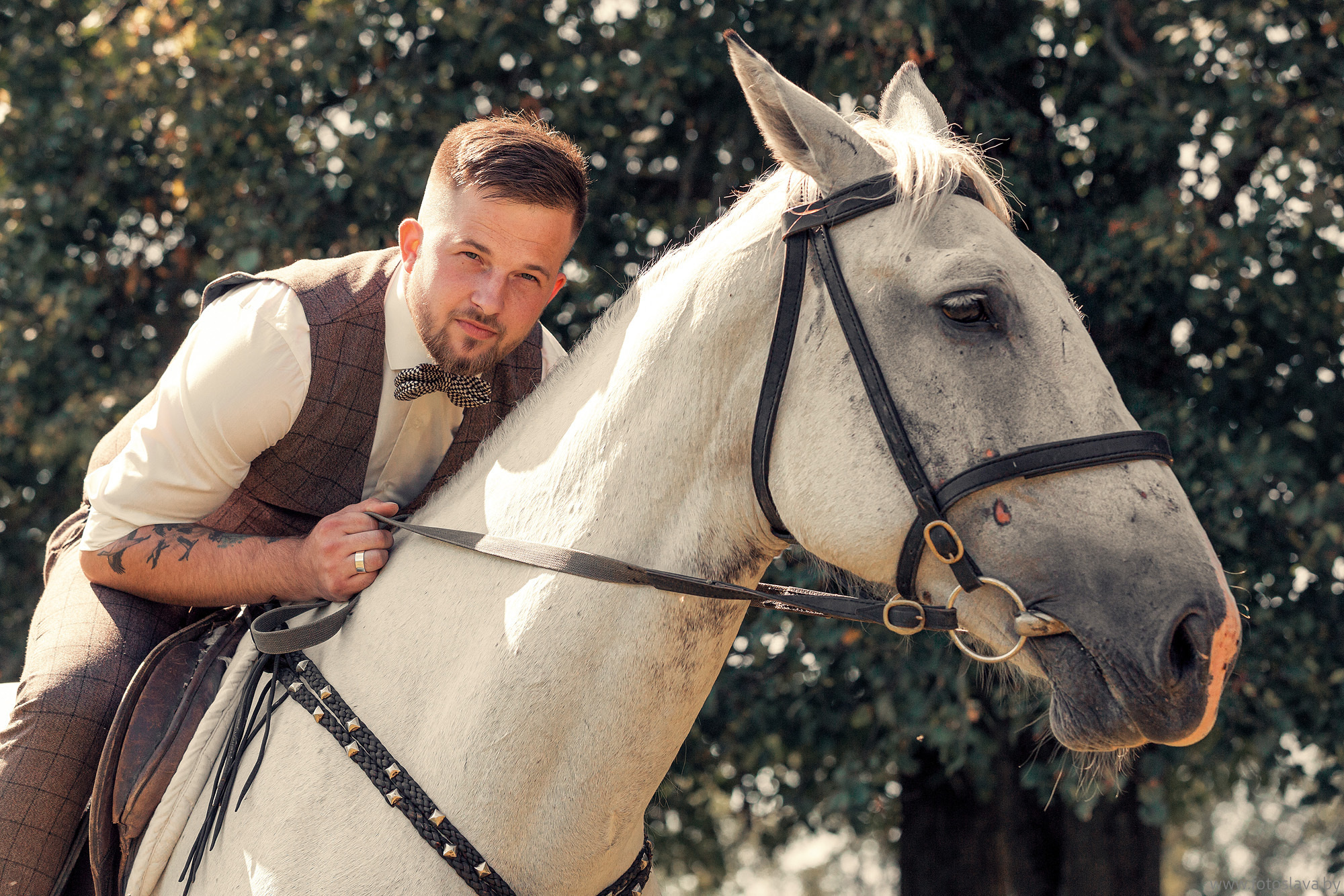 крупно жених сидит на белой лошади, прислонившись к ее шее.
