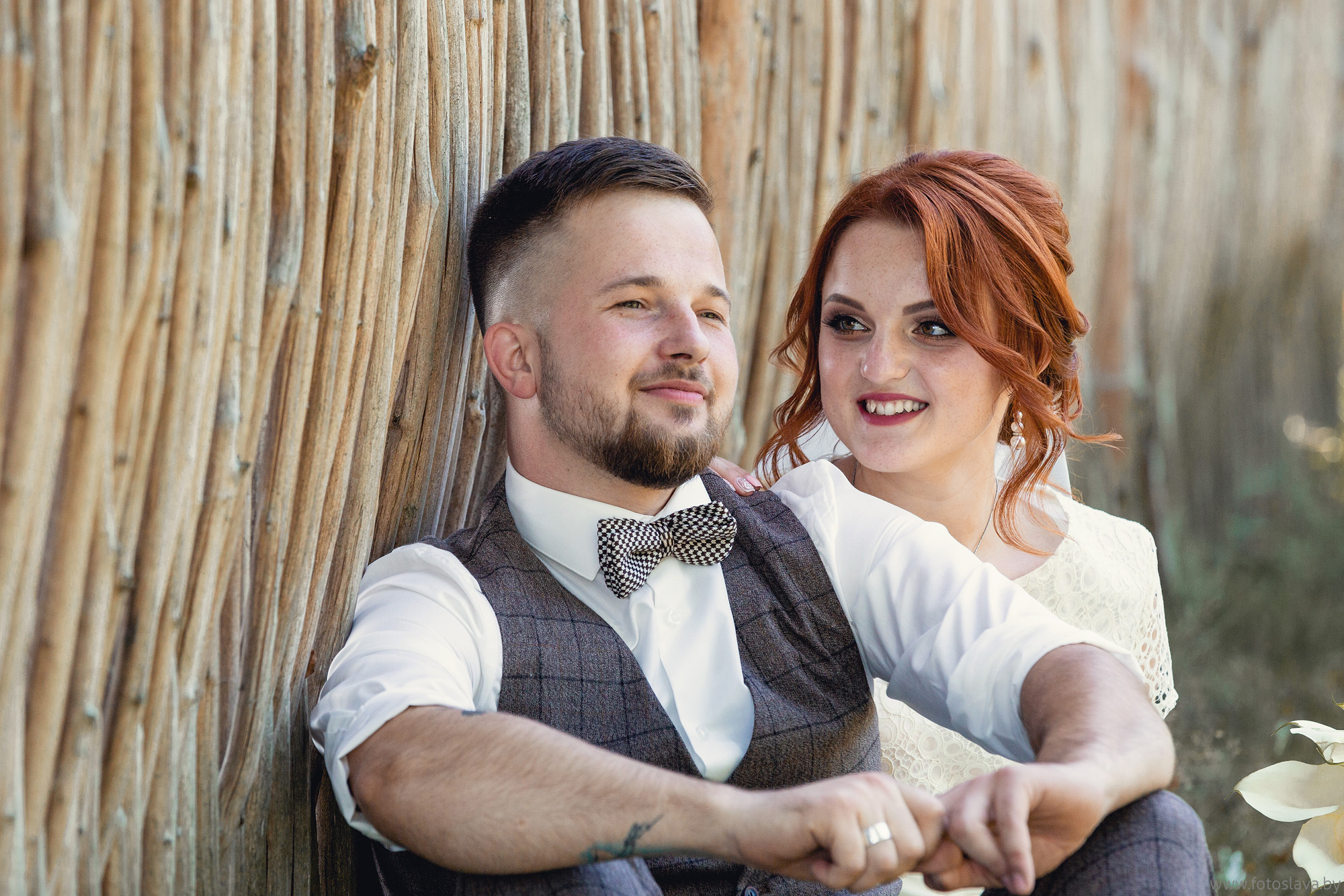 жених с невестой сидят на земле прислонившись к камышевому забору.