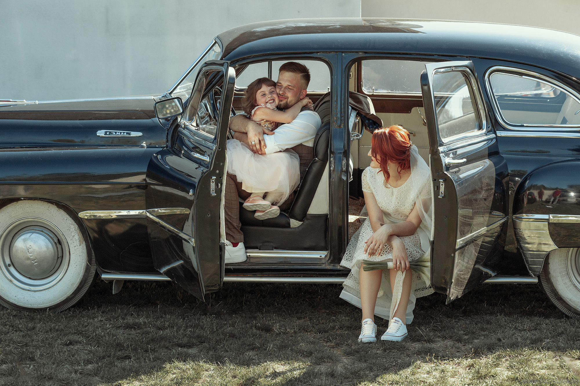 жених сидит в машине и обнимает ребенка, невеста рядом смотрит на них.