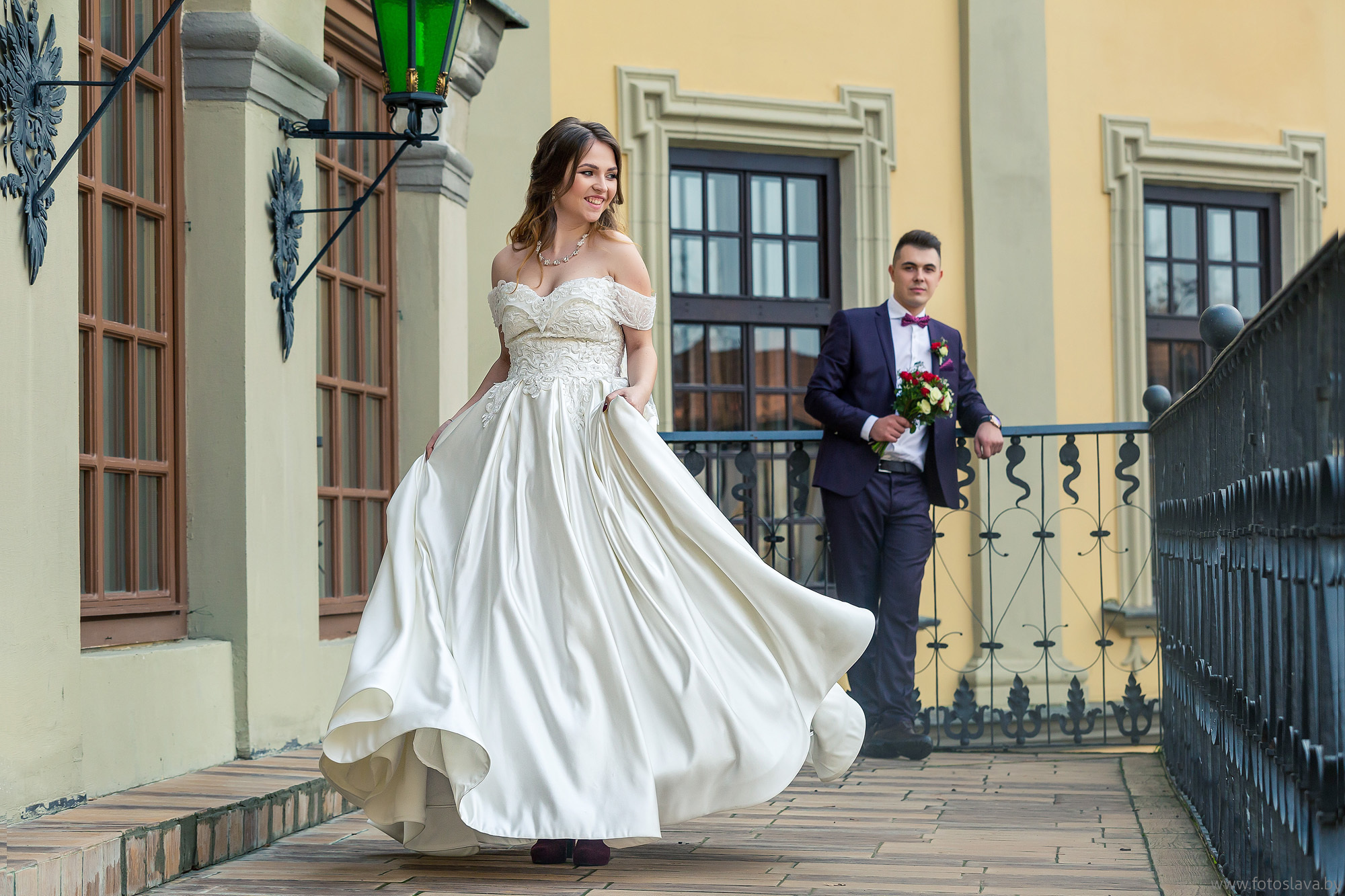 невеста в полный рост на фоне жениха на балконе замка.