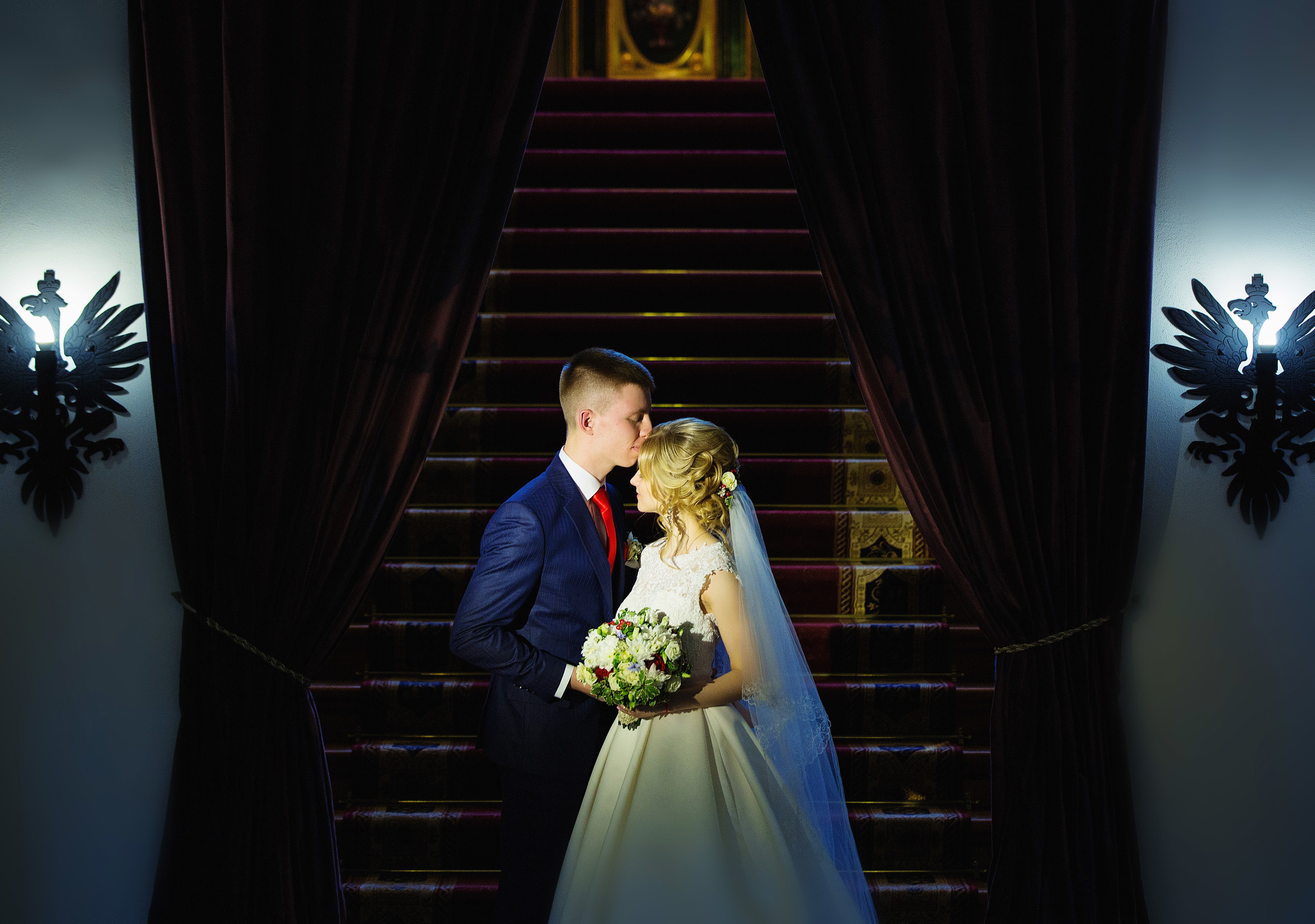 жених целует невесту в лоб на фоне парадной лестницы.