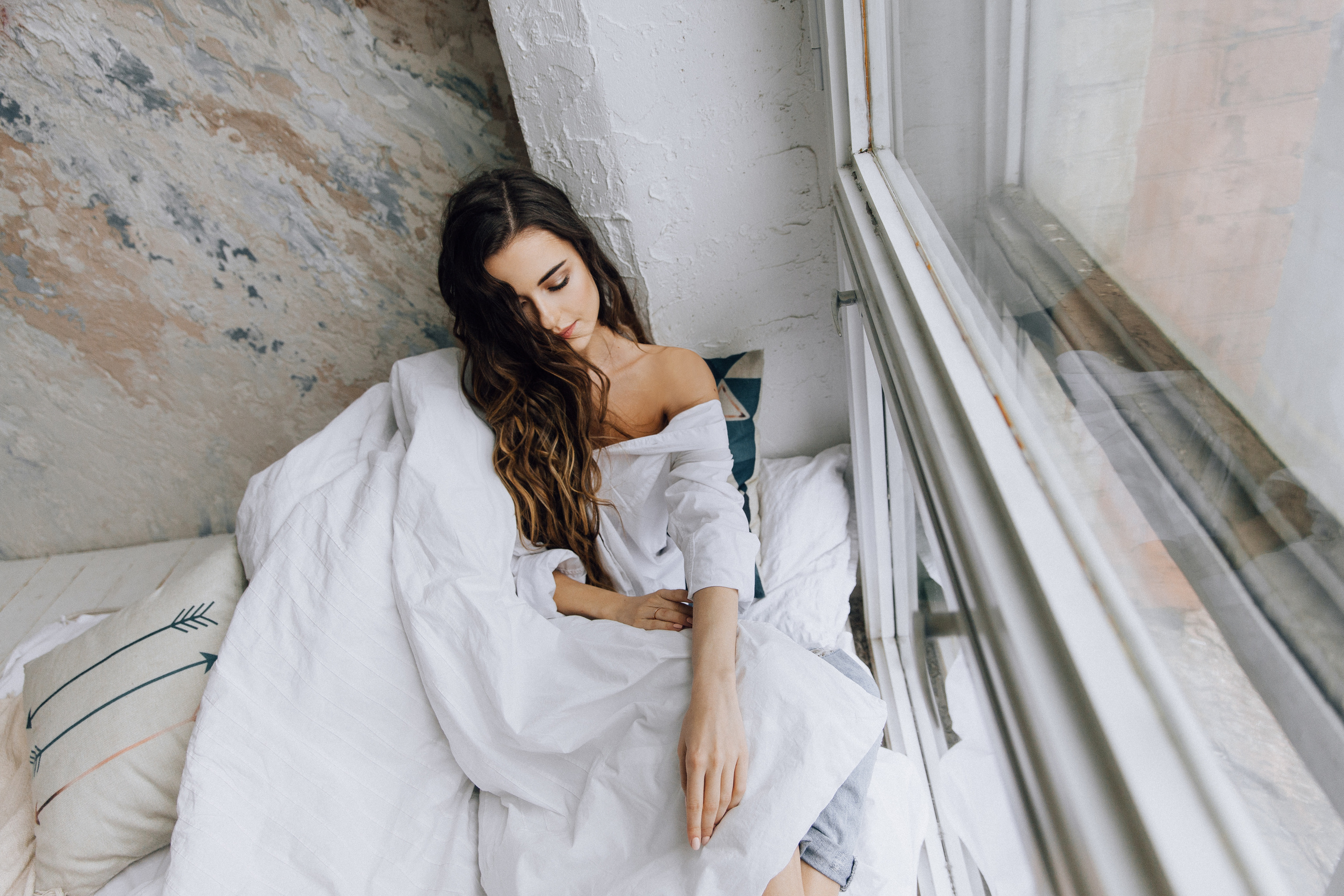 Девушка с длинными волосами в белой рубашке сидит на кровати у окна