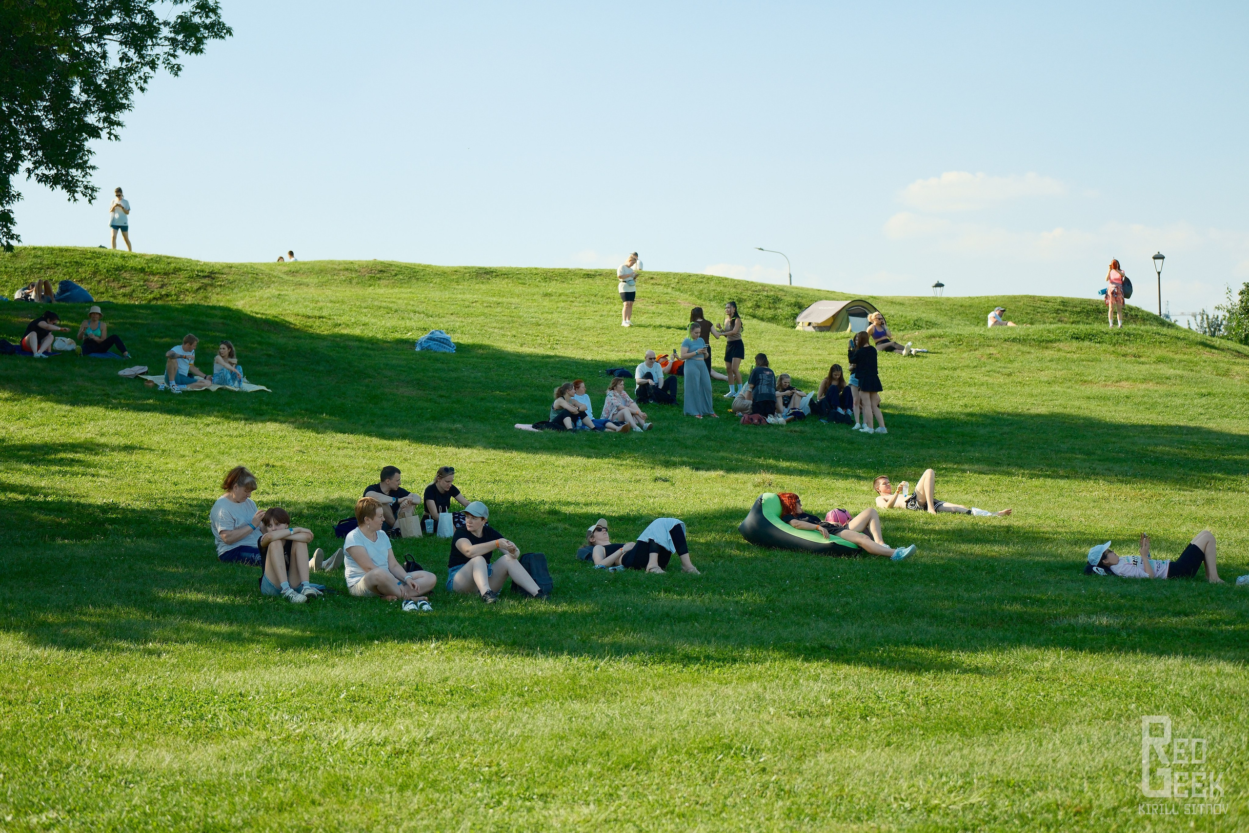 Люди отдыхают на зеленой поляне, слушая концерт.