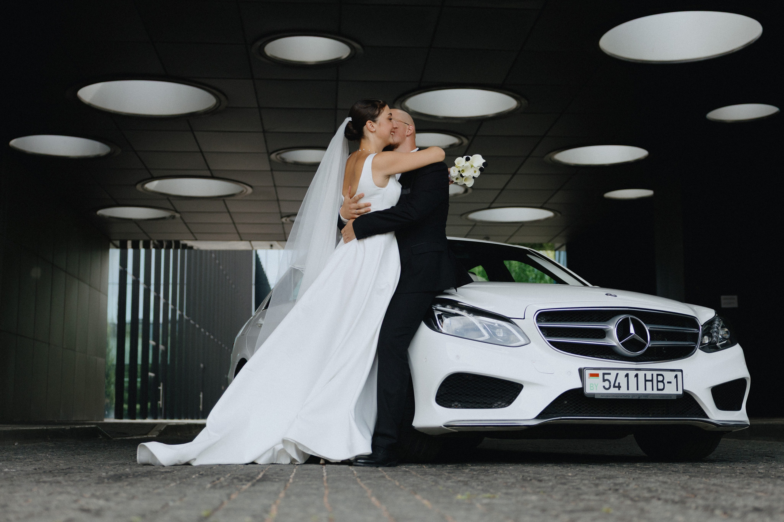 Жених и невеста позируют рядом с белым автомобилем Mercedes у Хилтон отеля.
