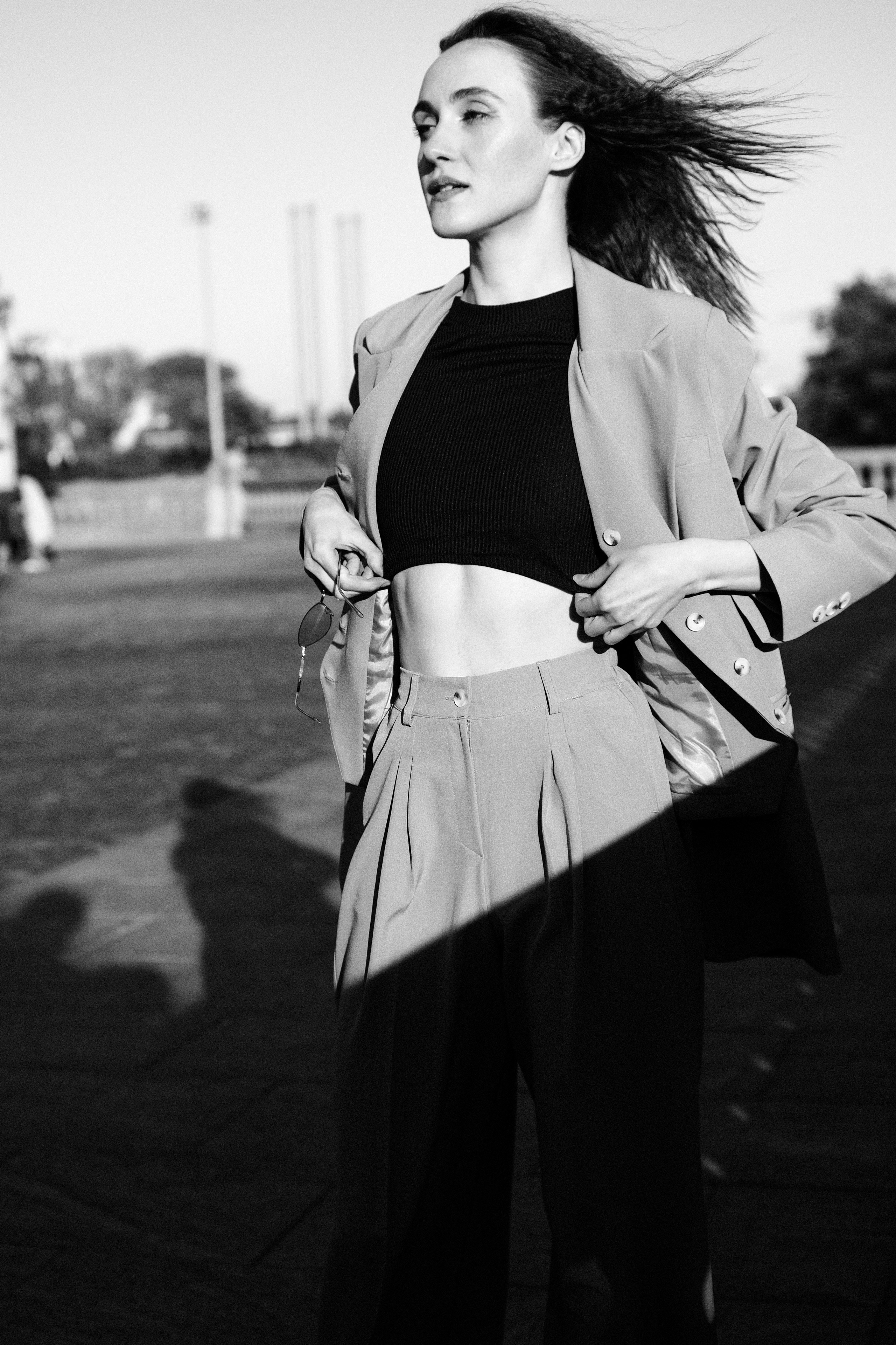 Черно-белый уличный портрет девушки в бежевом костюме с черным кроп-топом, развевающиеся на ветру волосы, динамичная поза, солнечный свет