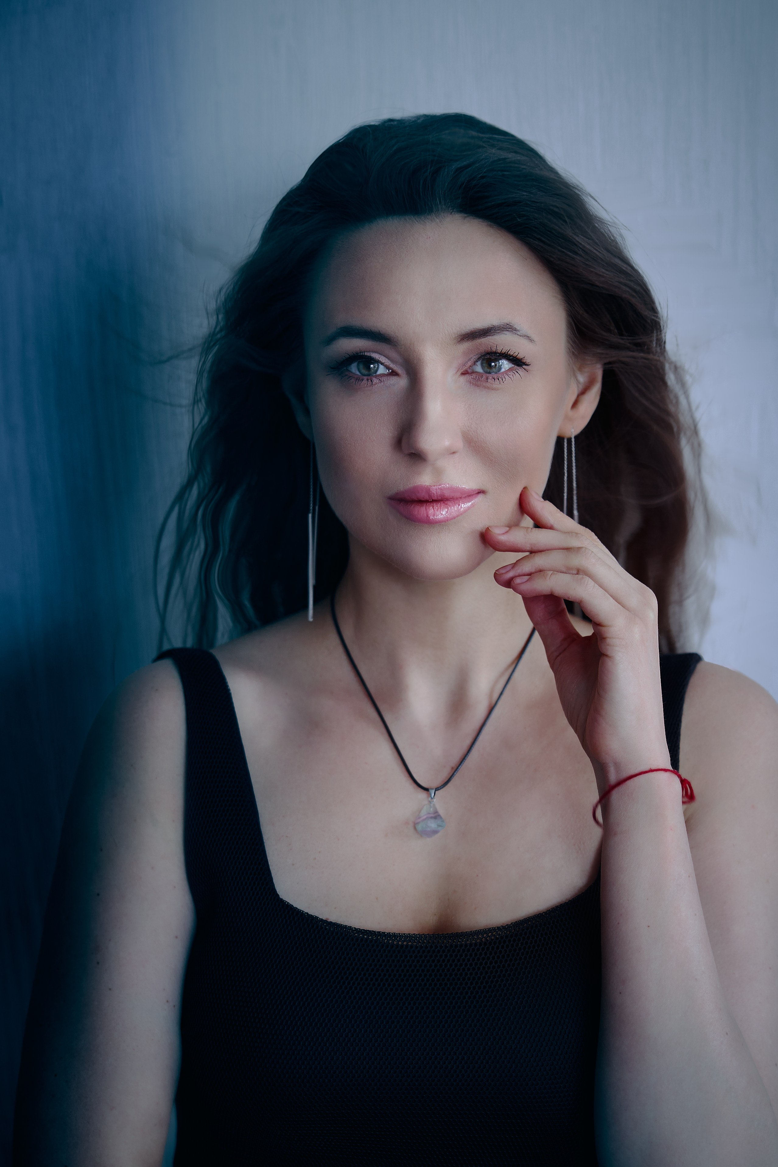 Портрет крупным планом женщины с длинными каштановыми волосами, в чёрном топе и с небольшим кулоном на шее. На губах у неё розовая помада, она нежно касается подбородка, на запястье тонкая красная нить. Фотография выполнена в прохладном, мягком голубом свете на текстурированном белом фоне.