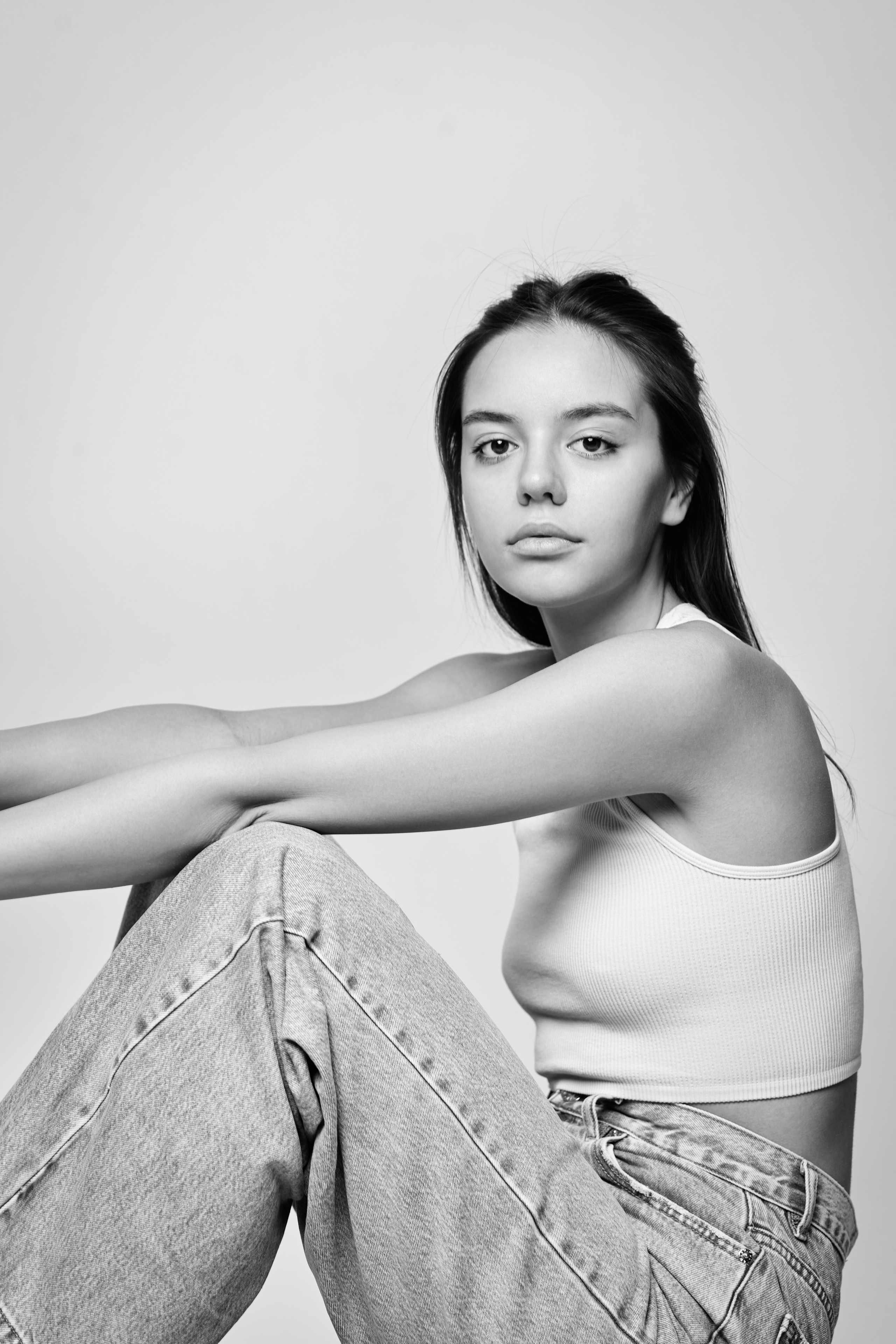Сексуальная темноволосая модель Амина на светлом фоне в белом топике и джинсах с высокой талией.