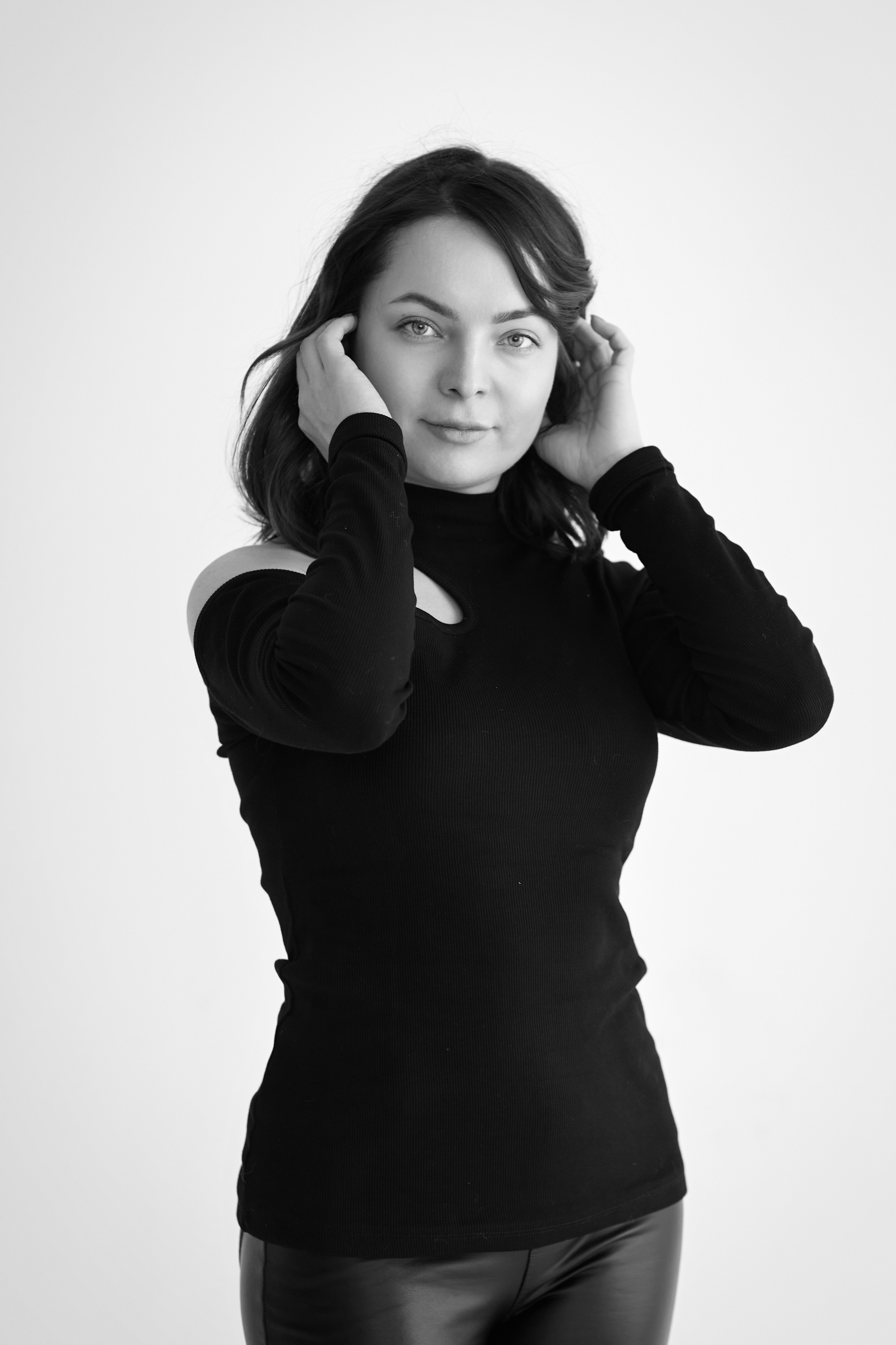 Темноволосая модель Татьяна на светлом фоне, в светлом платье, рубашке или топике или на стуле, смотрящая с легкой улыбкой.