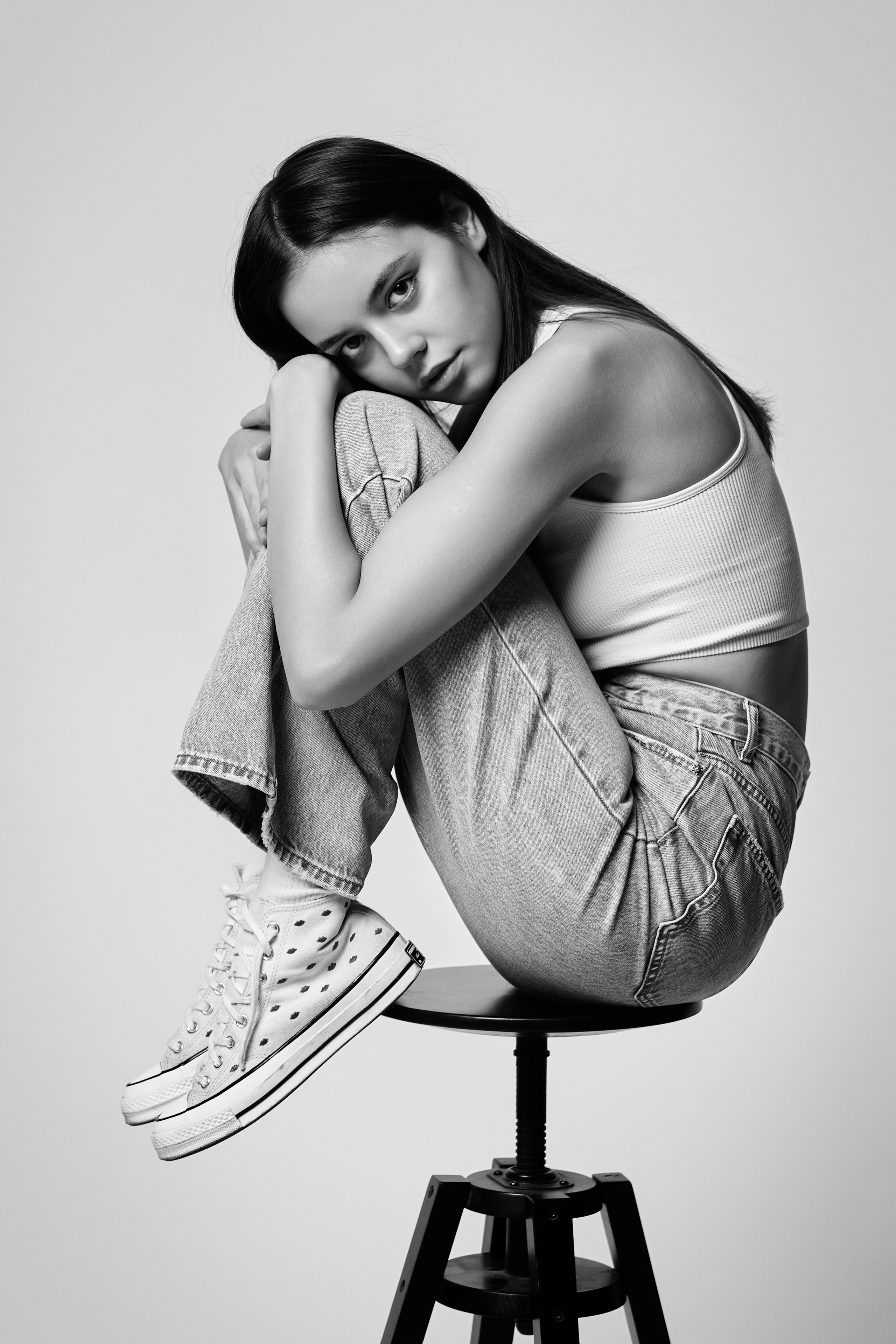 Модель Амина стоит в студии на светлом фоне, одетая в светлое платье, ее темные волосы опускаются на плечи.